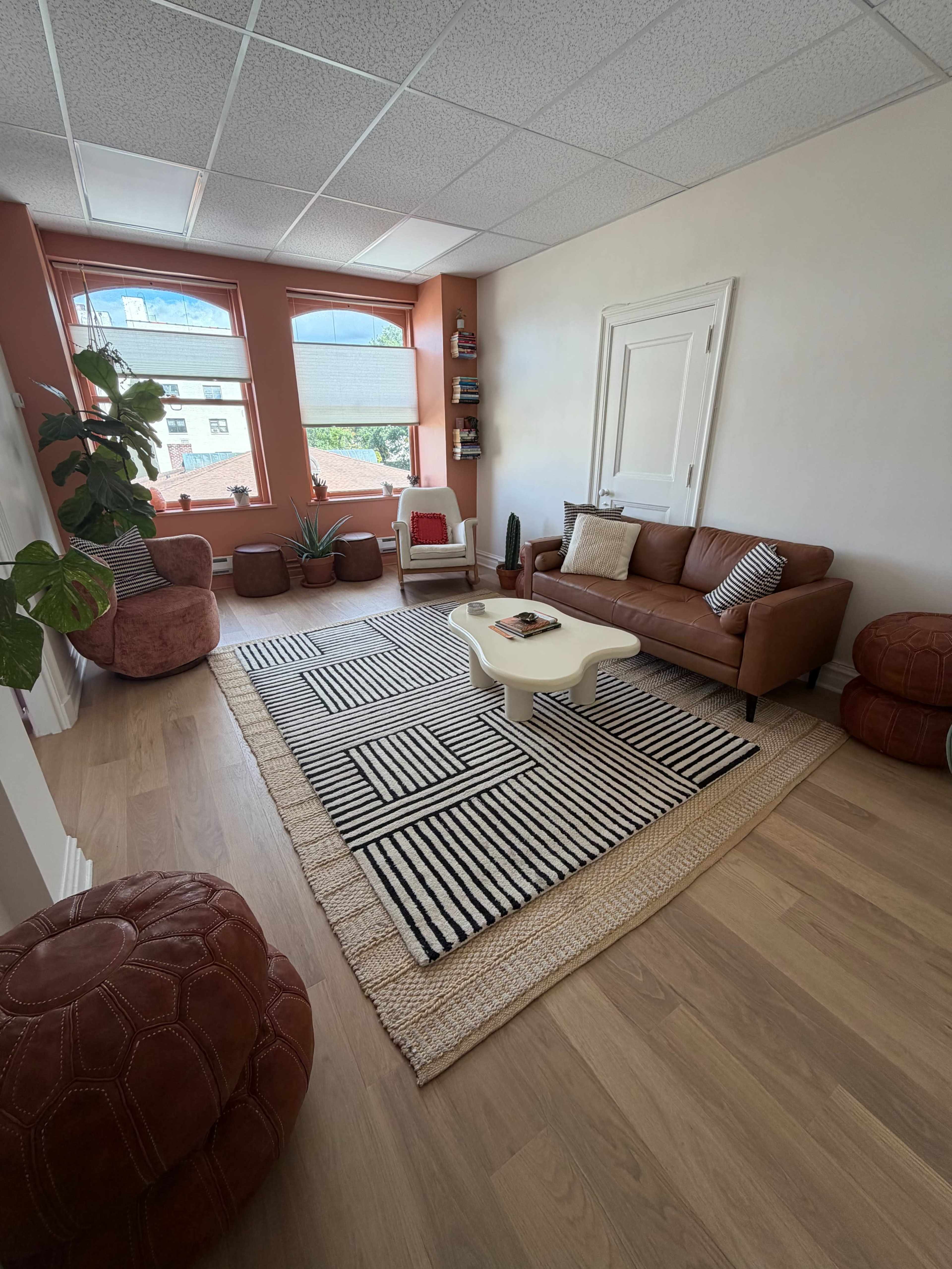 The image shows a cozy living room with a brown leather sofa, a white coffee table, and various plants arranged around a striped rug on wooden flooring.