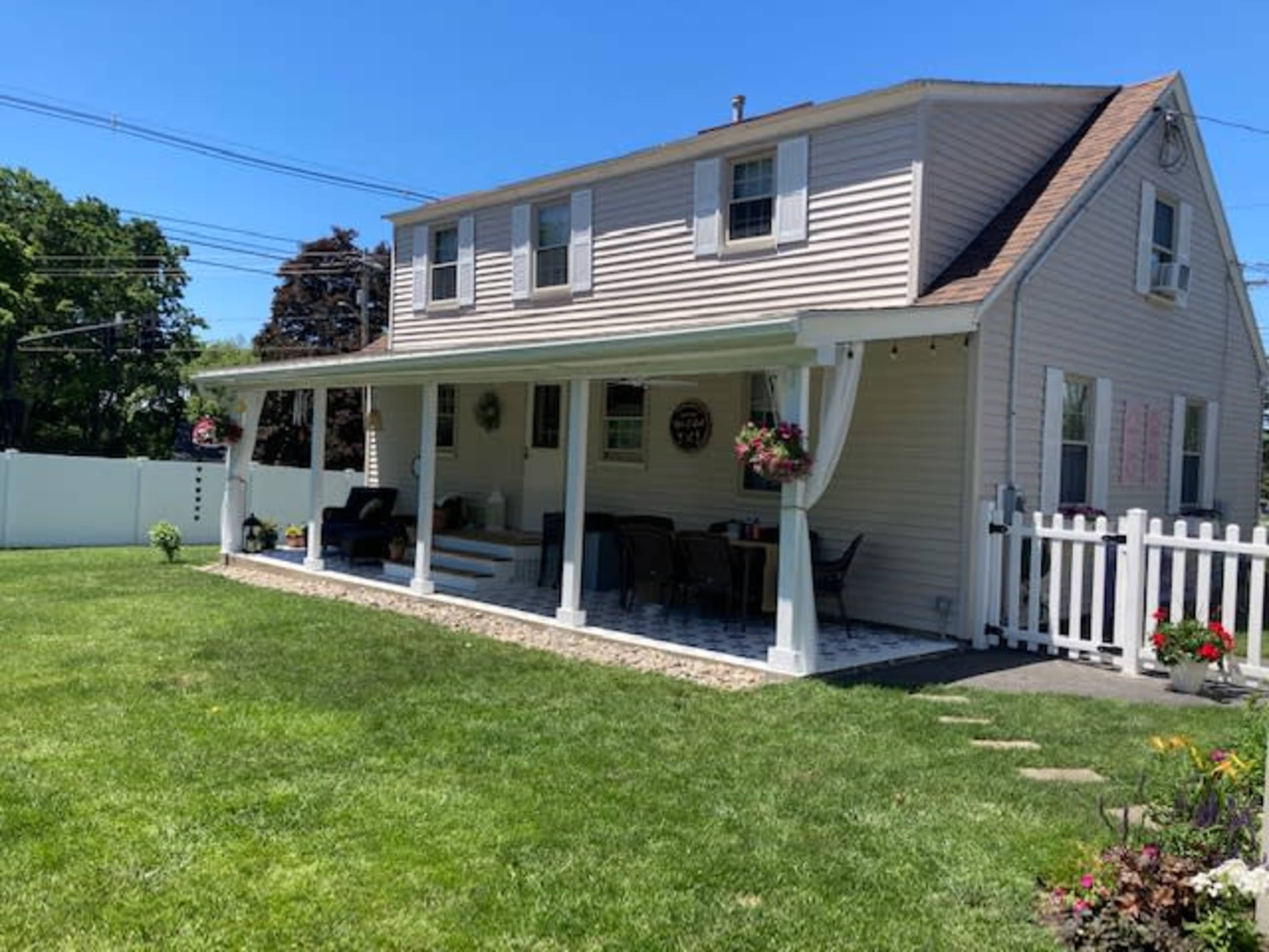 A two-story house with a porch, surrounded by a well-maintained lawn and a white picket fence, features outdoor seating and decorative planters.