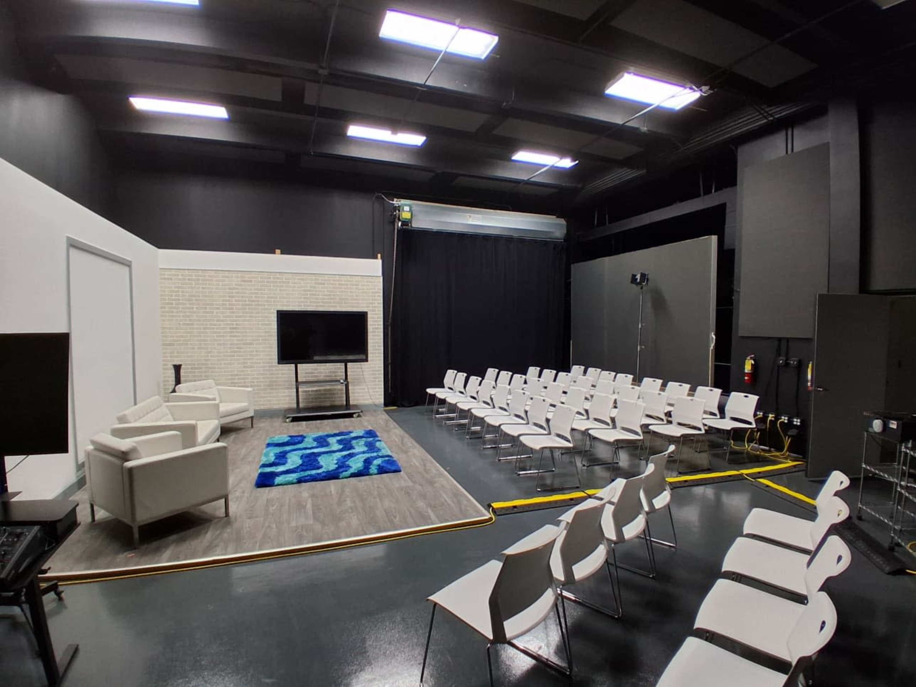 The image shows a modern studio space with white chairs arranged in rows, a blue rug on the floor, a television on a stand, and a pair of white armchairs against a white brick wall.
