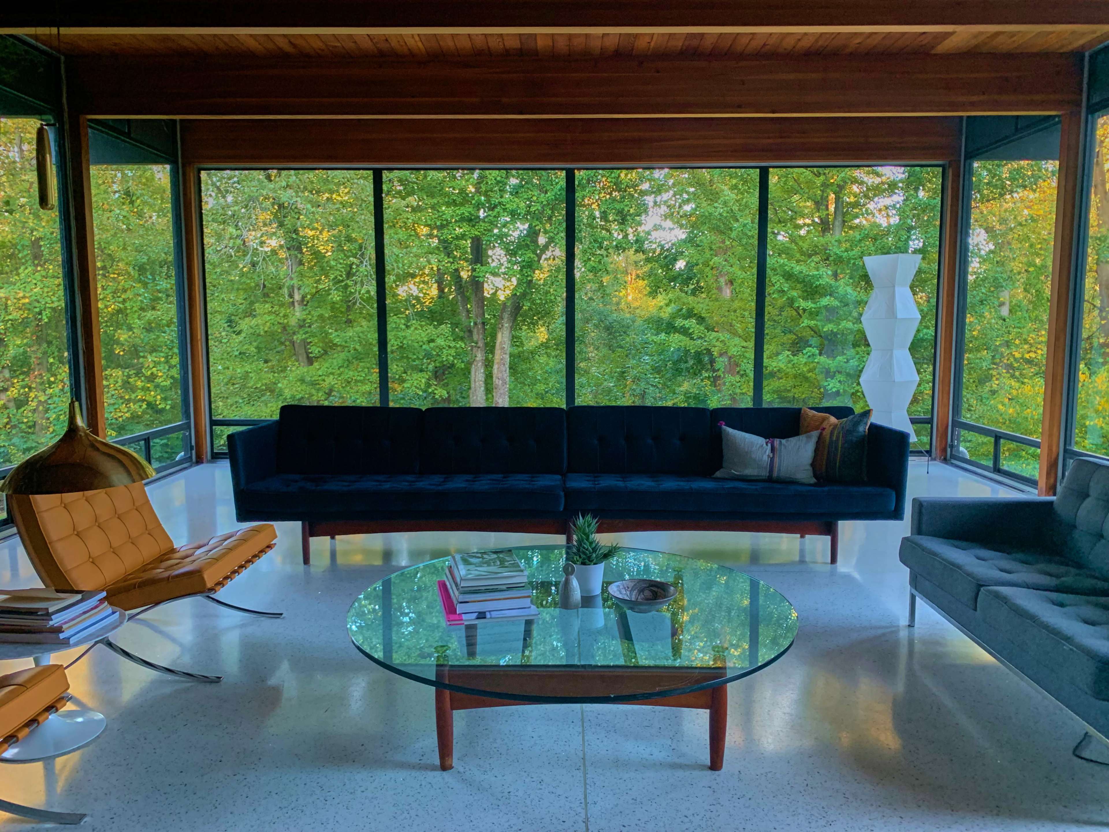 The image shows a modern living room with large windows overlooking a green wooded area, featuring a dark sofa, a glass coffee table, and two stylish chairs.