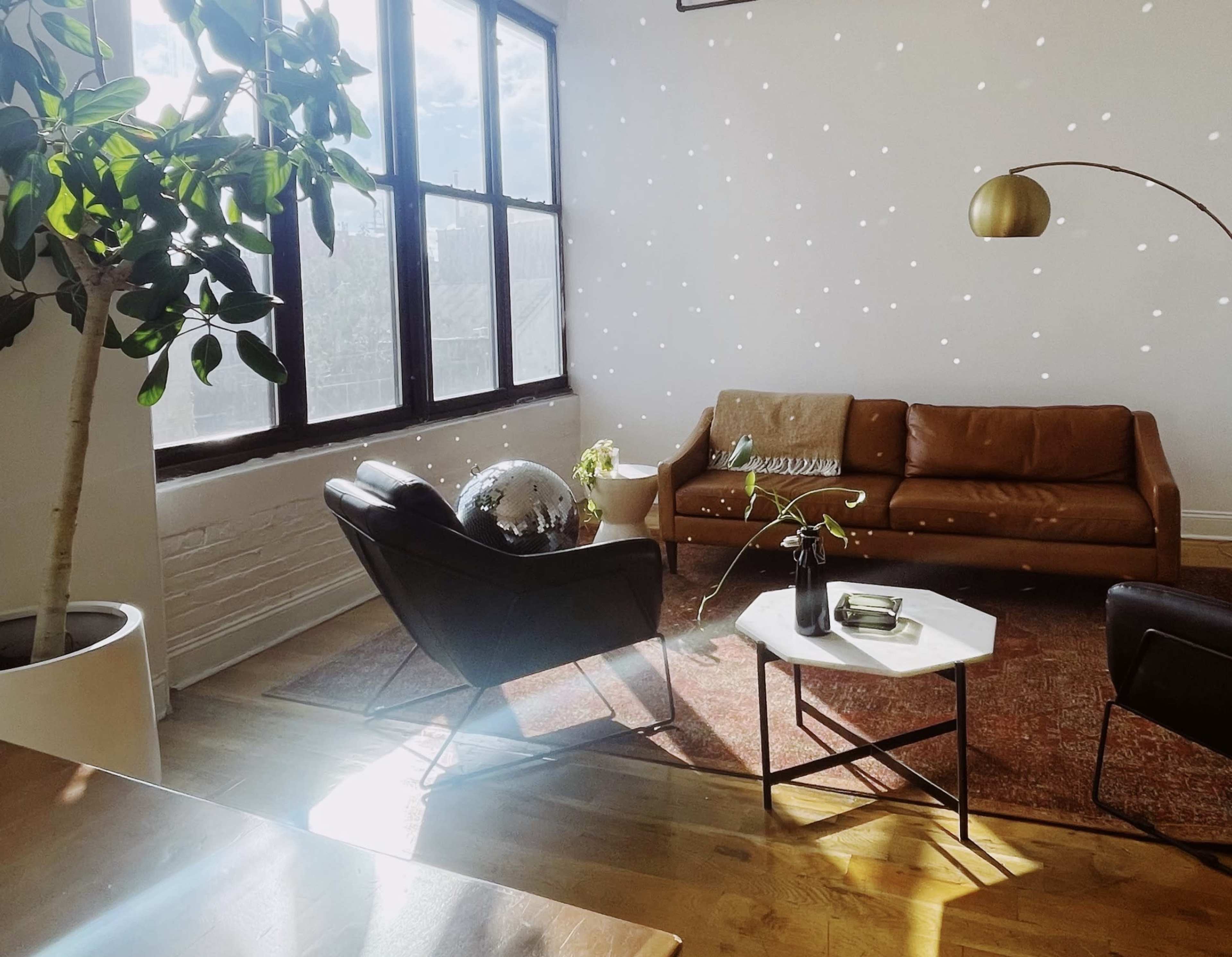 A bright living room features a brown leather sofa, two black chairs, a round coffee table, and a large window with sunlight creating scattered reflections on the walls.