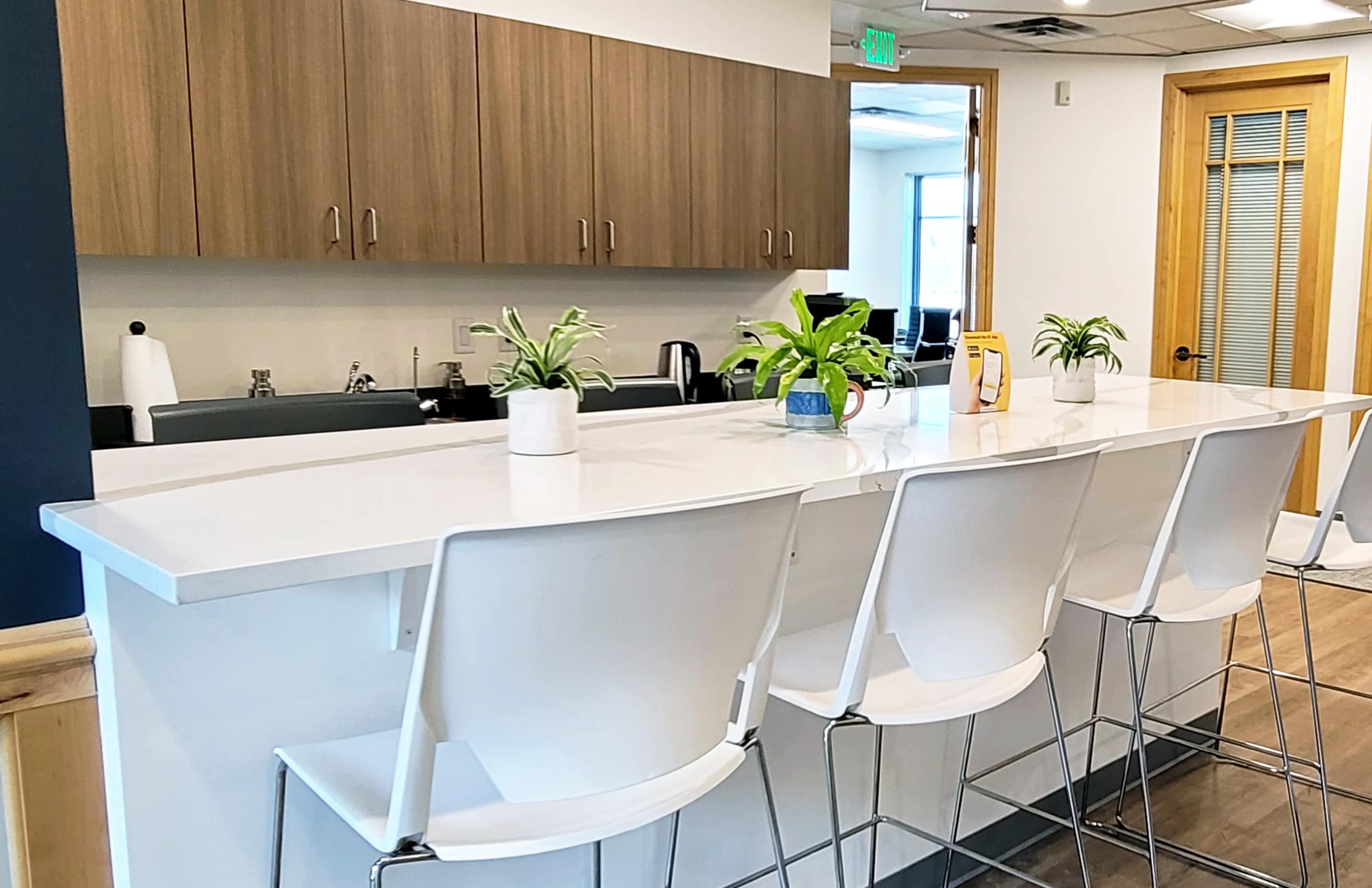 A modern kitchen features a white countertop bar with four white stools and potted plants on the surface.