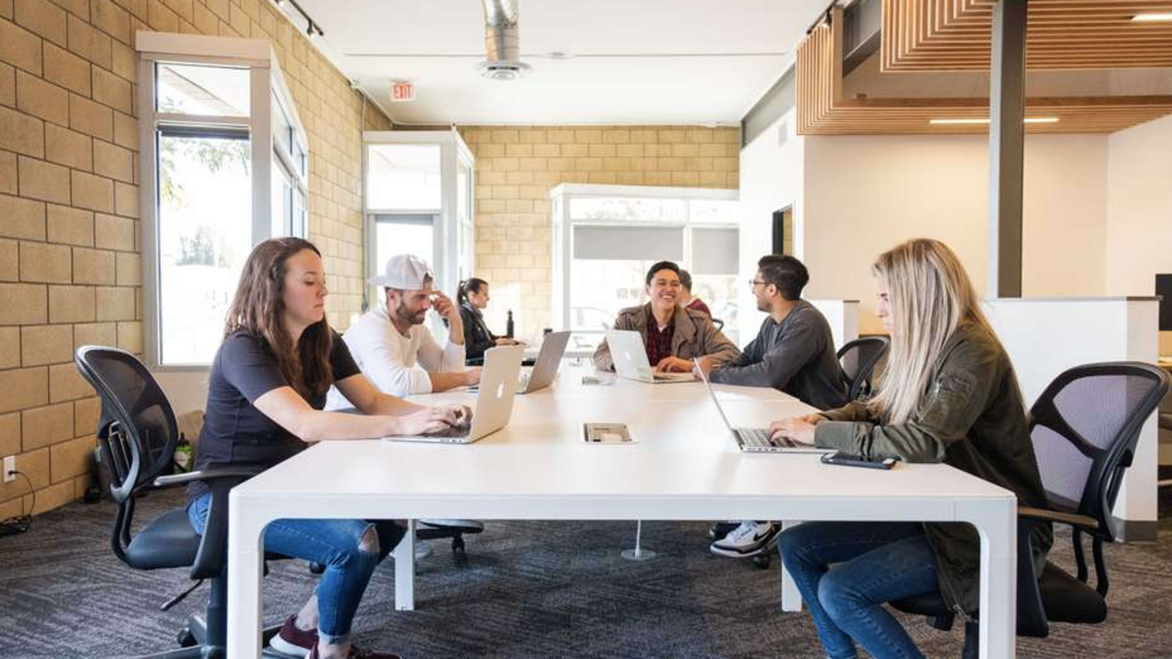 A group of six people is working on laptops at a large white table in a modern office space with brick walls.