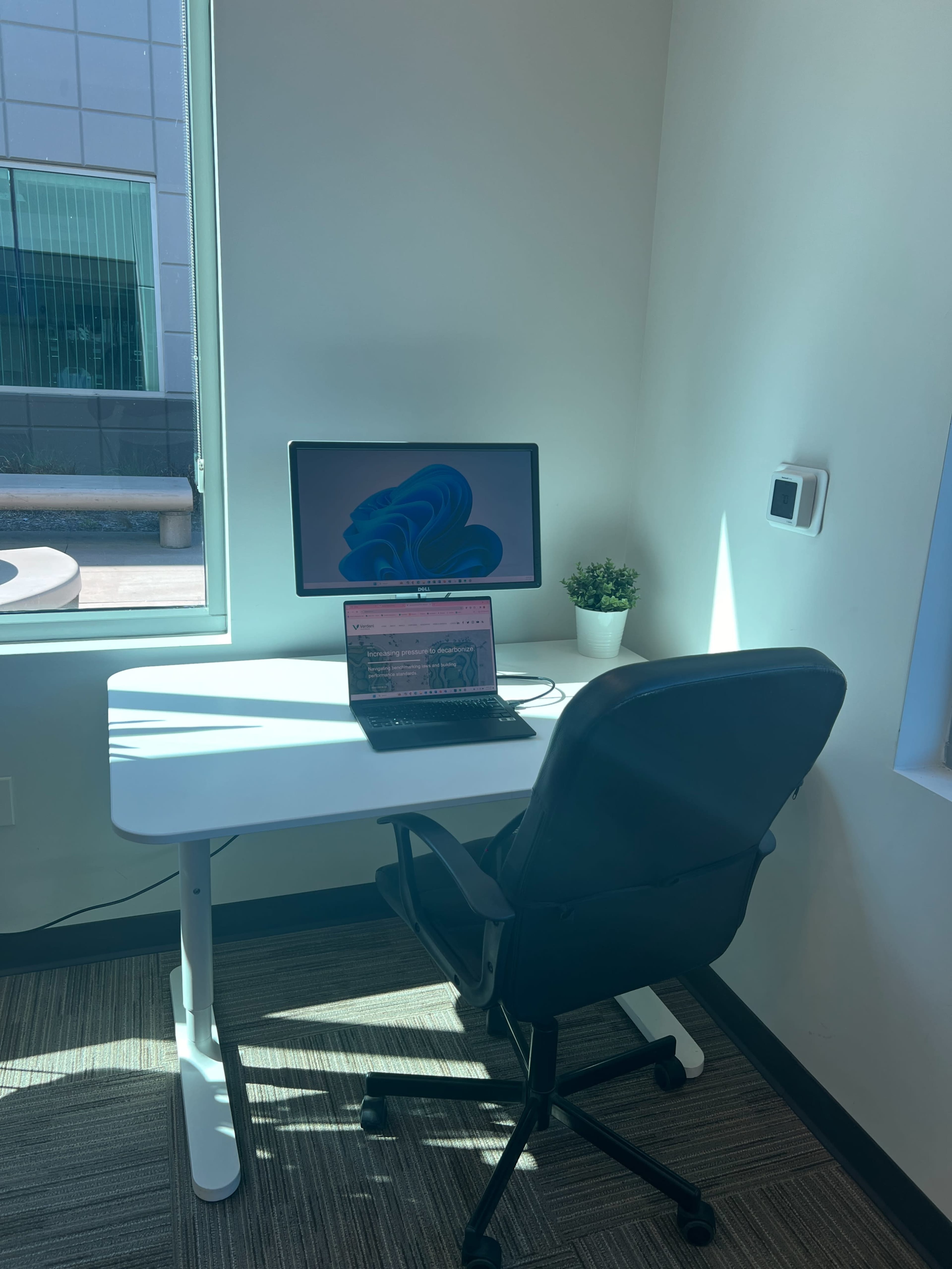 A modern workspace features a desk with a monitor and a laptop, accompanied by a small plant, in a well-lit corner with a window.