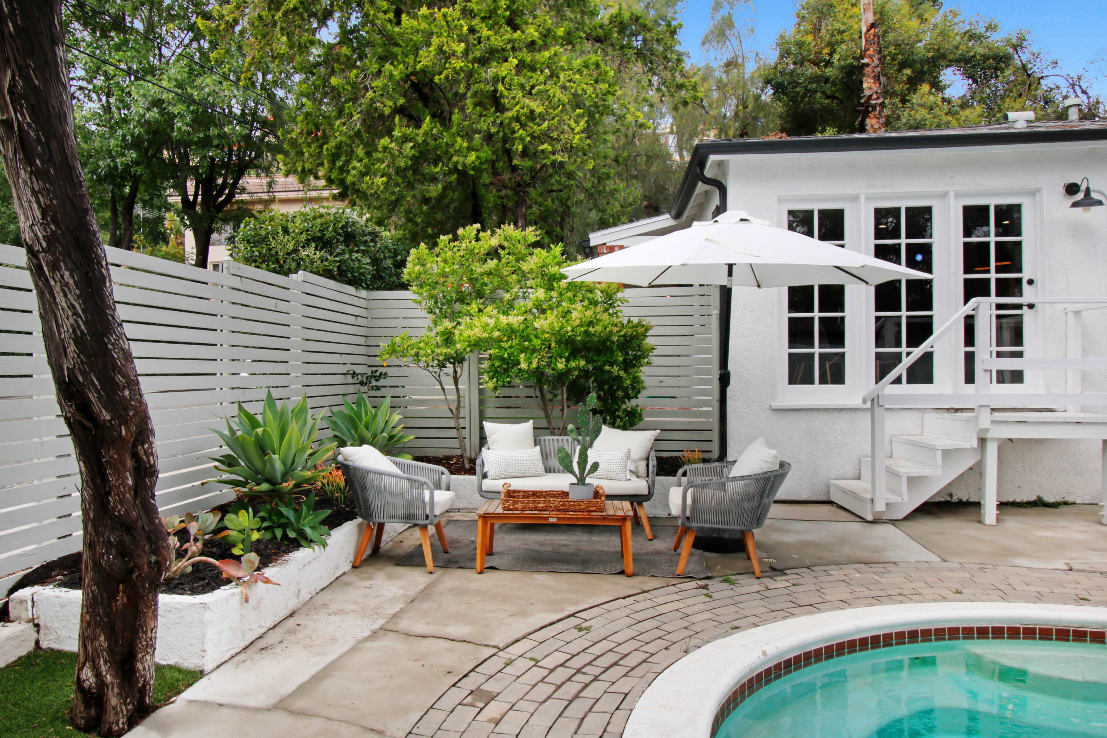 A cozy outdoor seating area features two chairs and a table under a white umbrella, adjacent to a pool and surrounded by plants and a white fence.
