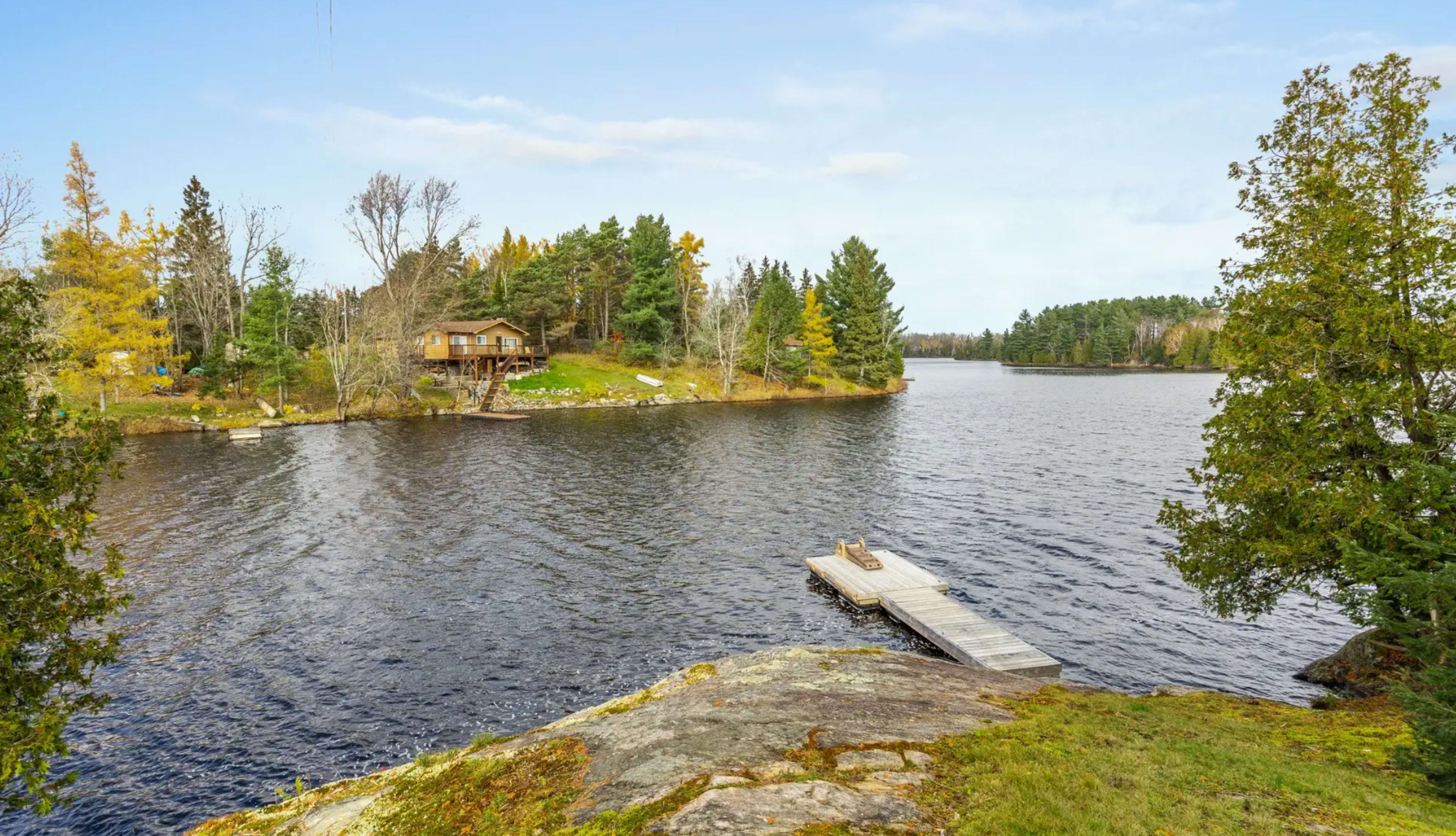 A calm lake with a wooden dock extending into the water, surrounded by trees and a cabin on the opposite shore.