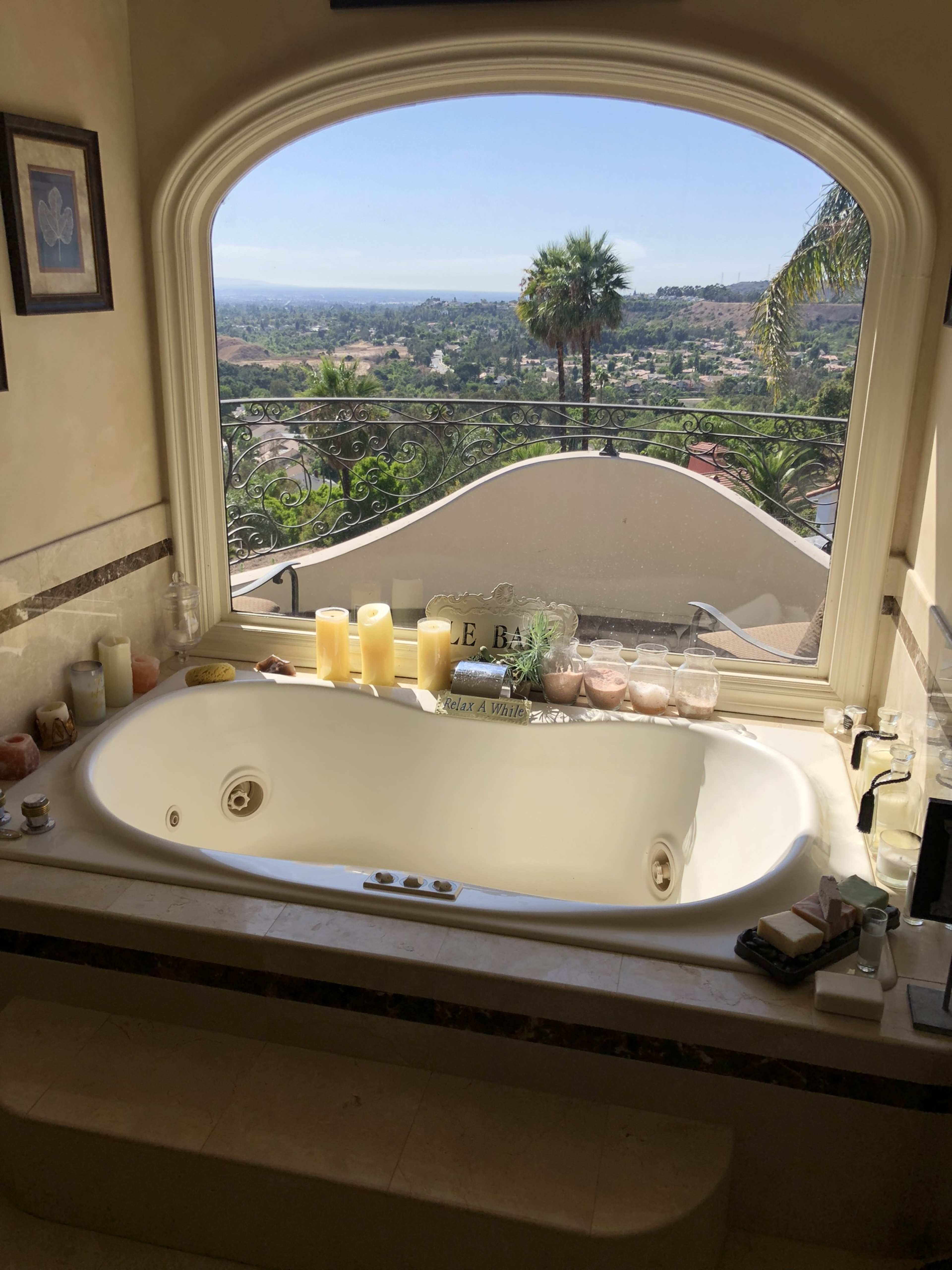 A large jacuzzi tub is positioned beneath a wide arched window that offers a view of a landscape filled with trees and distant hills.