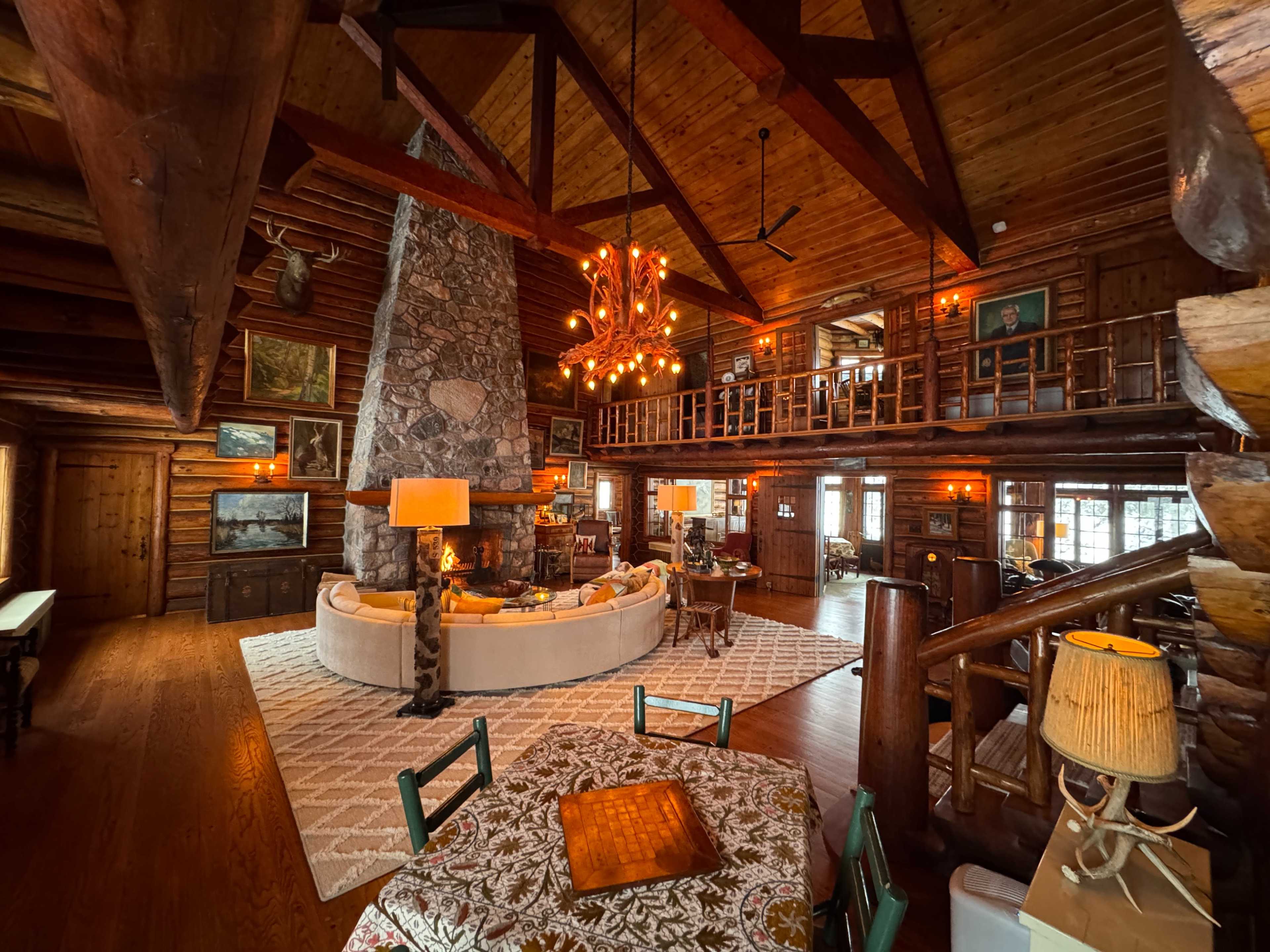 The image shows a spacious log cabin interior with a large stone fireplace, wooden beams, and multiple seating areas.