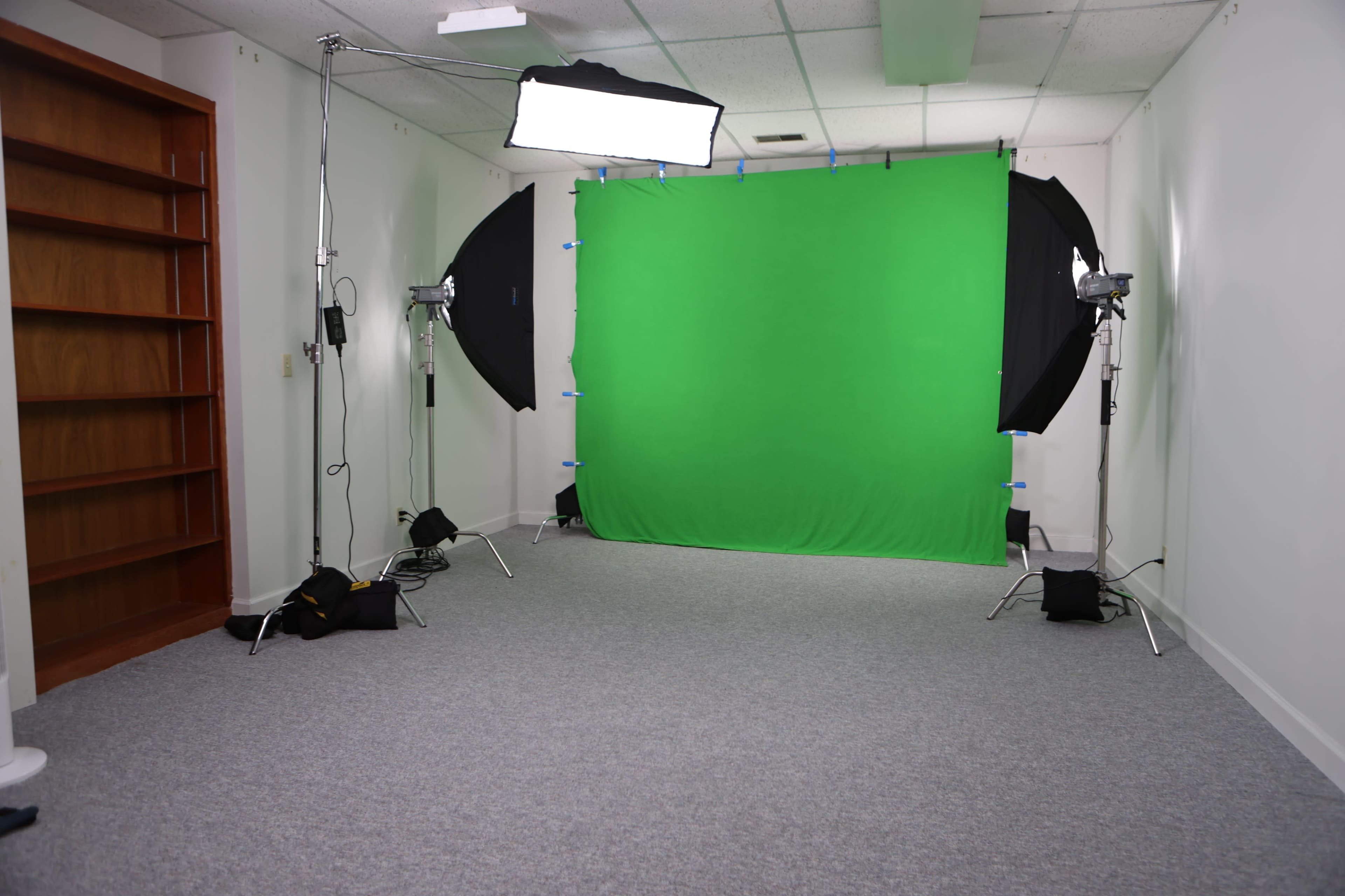 The image shows a studio setup featuring a green screen, lighting equipment, and a carpeted floor, with an empty bookshelf against one wall.