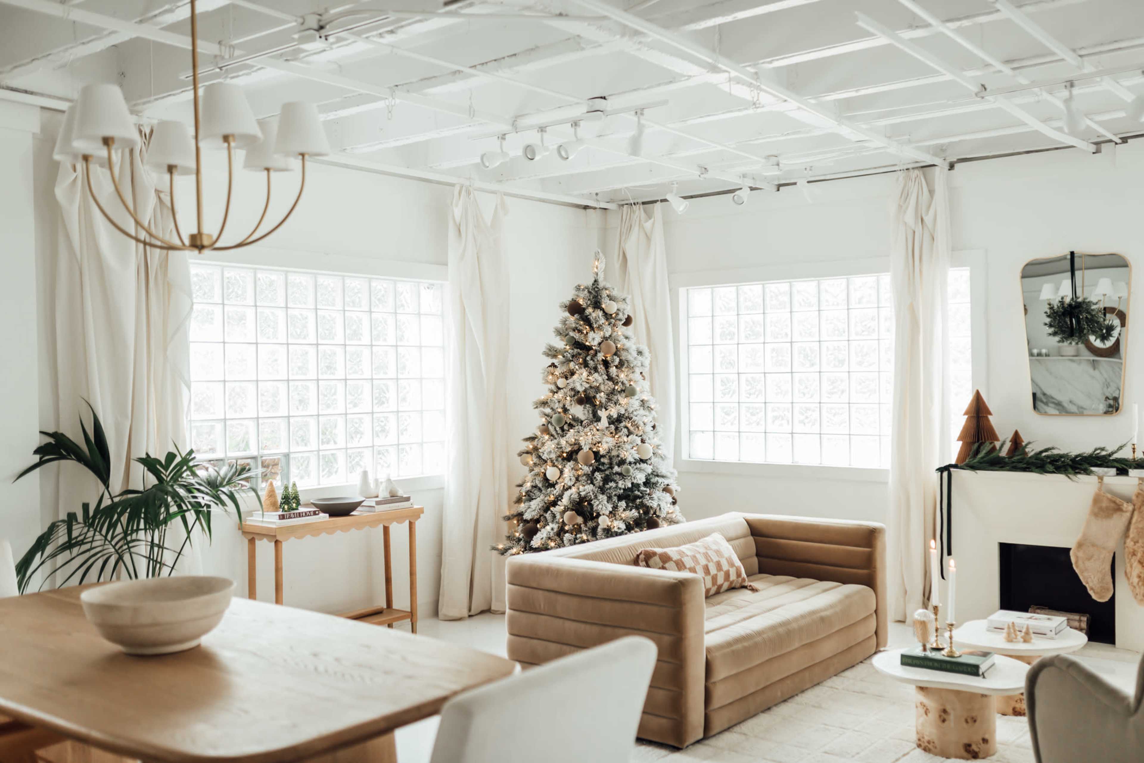 A cozy living room features a decorated Christmas tree, large windows, and a beige sofa, with a dining table and greenery accents nearby.