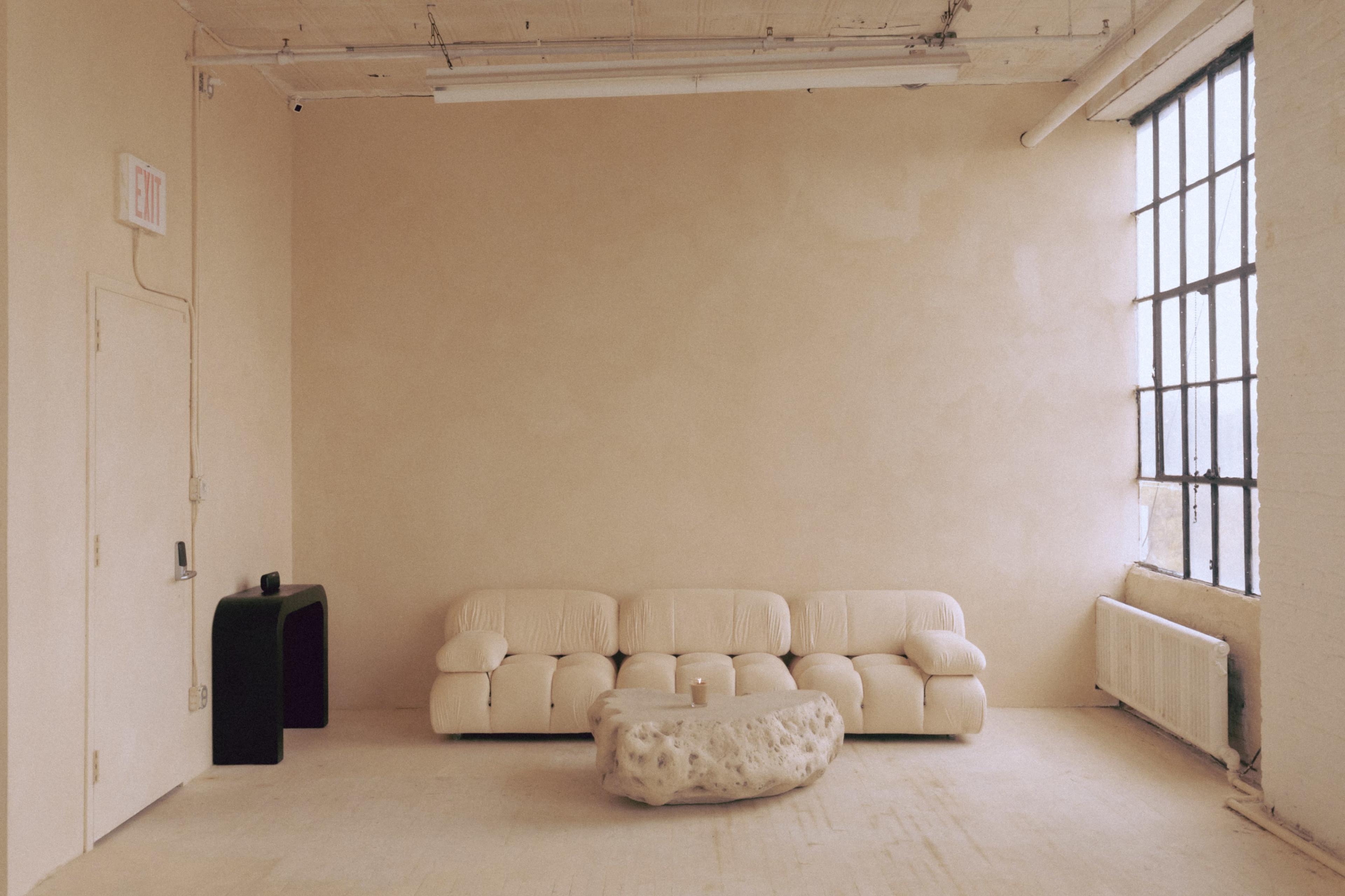 A minimalist interior features a light-colored couch, a rounded coffee table, and a large window with natural light.
