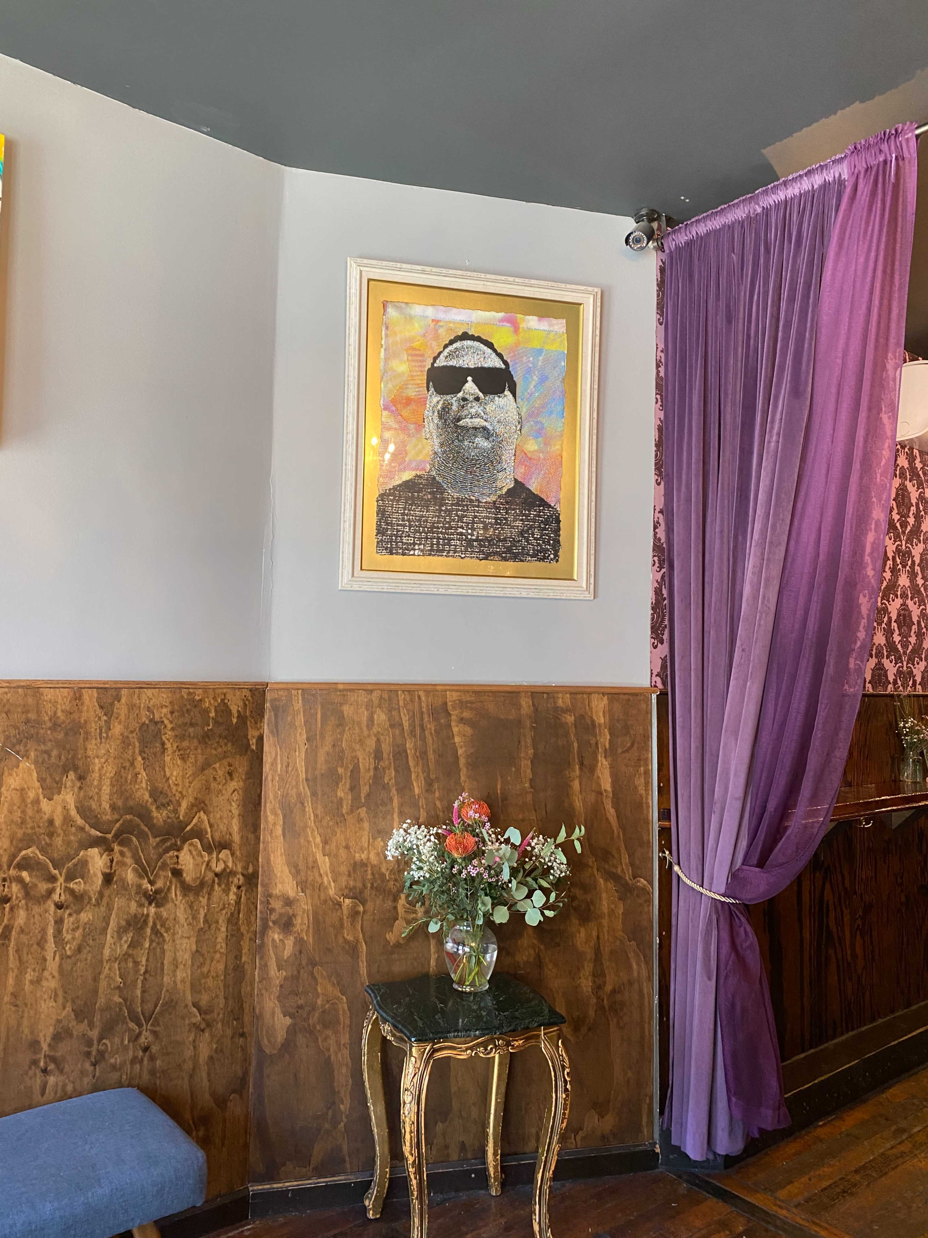 The image shows a framed portrait of a man wearing sunglasses hanging on a wall beside a purple curtain, with a small table and a vase of flowers in the foreground.