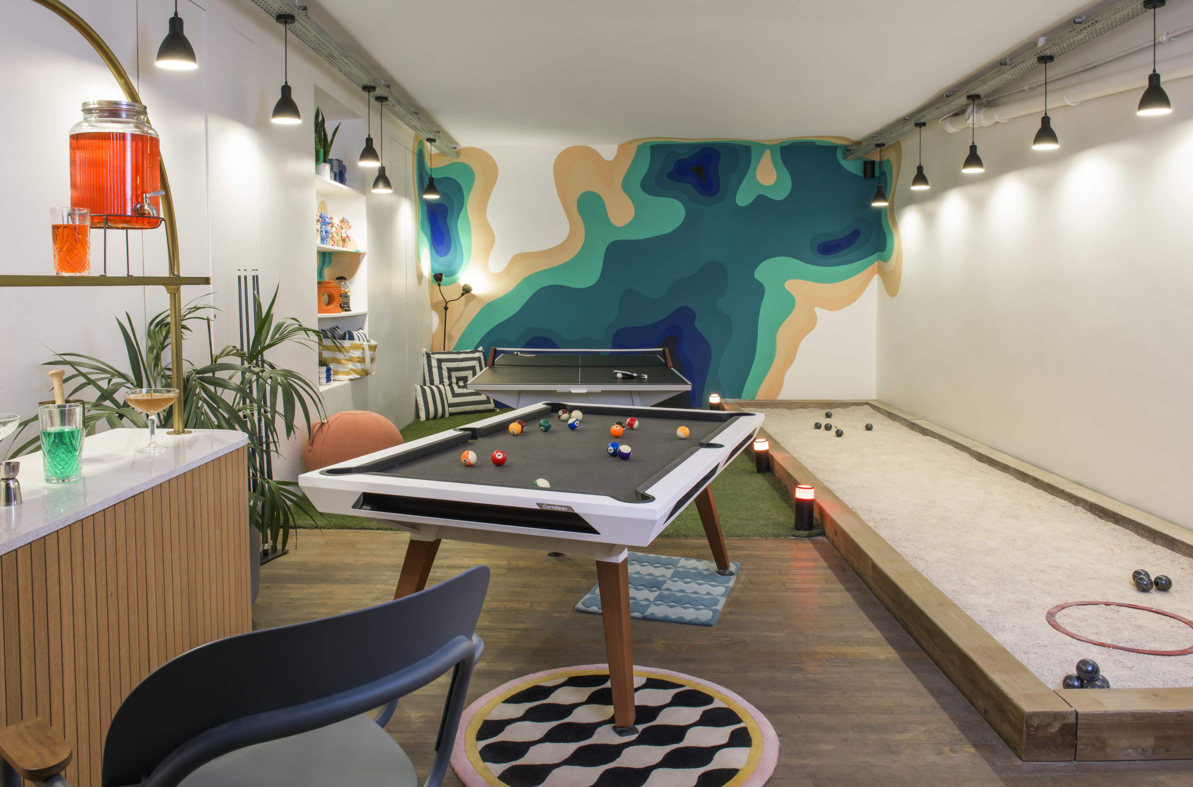 The image shows a game room featuring a modern pool table, a dining area with drinks displayed, and a decorative wall mural in vibrant colors.