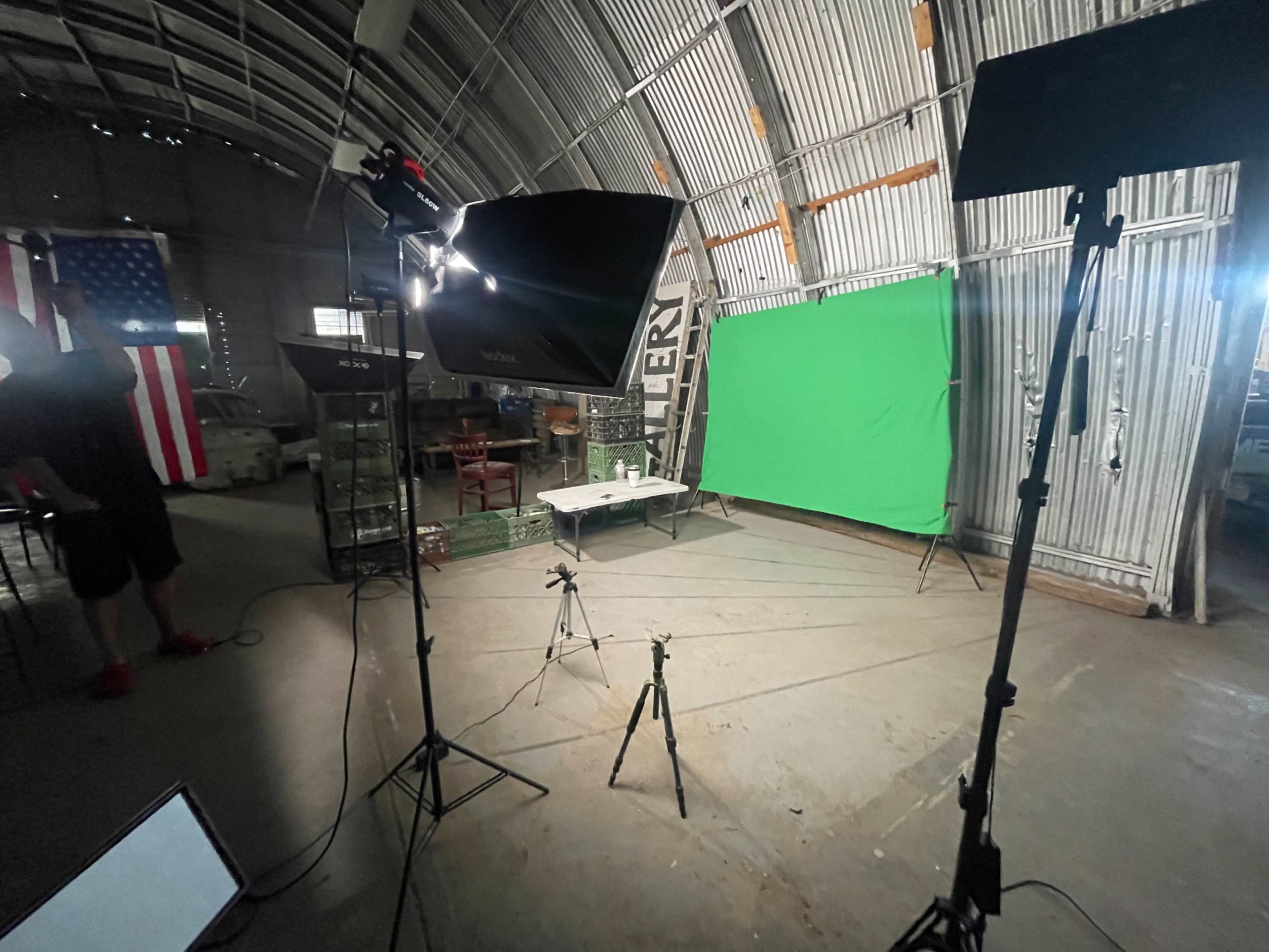 The image shows a studio setup in a large barn-like space, featuring a green screen, professional lighting equipment, and a nearby American flag.