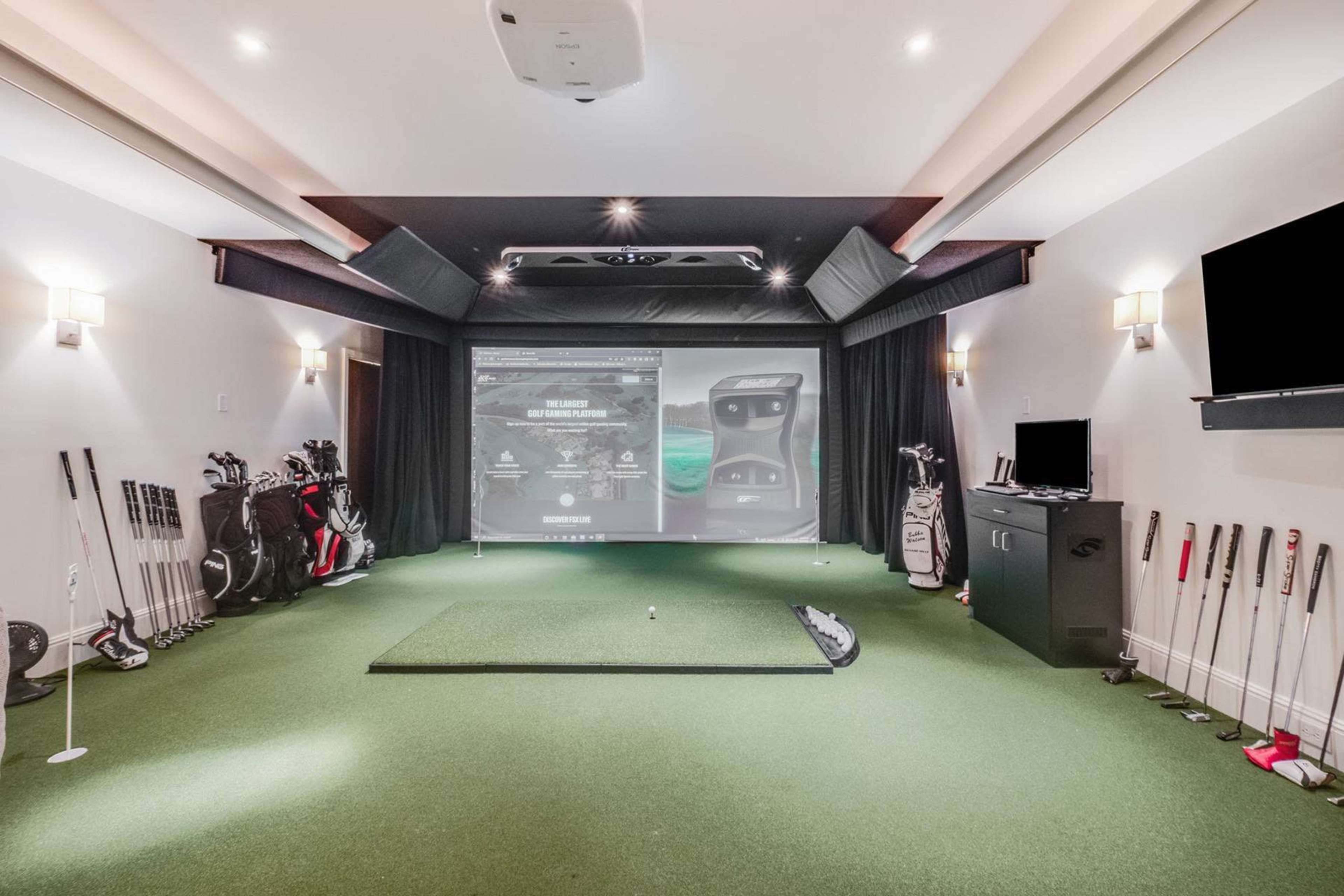 A home golf simulator setup featuring a putting green, golf clubs, and a large projection screen.