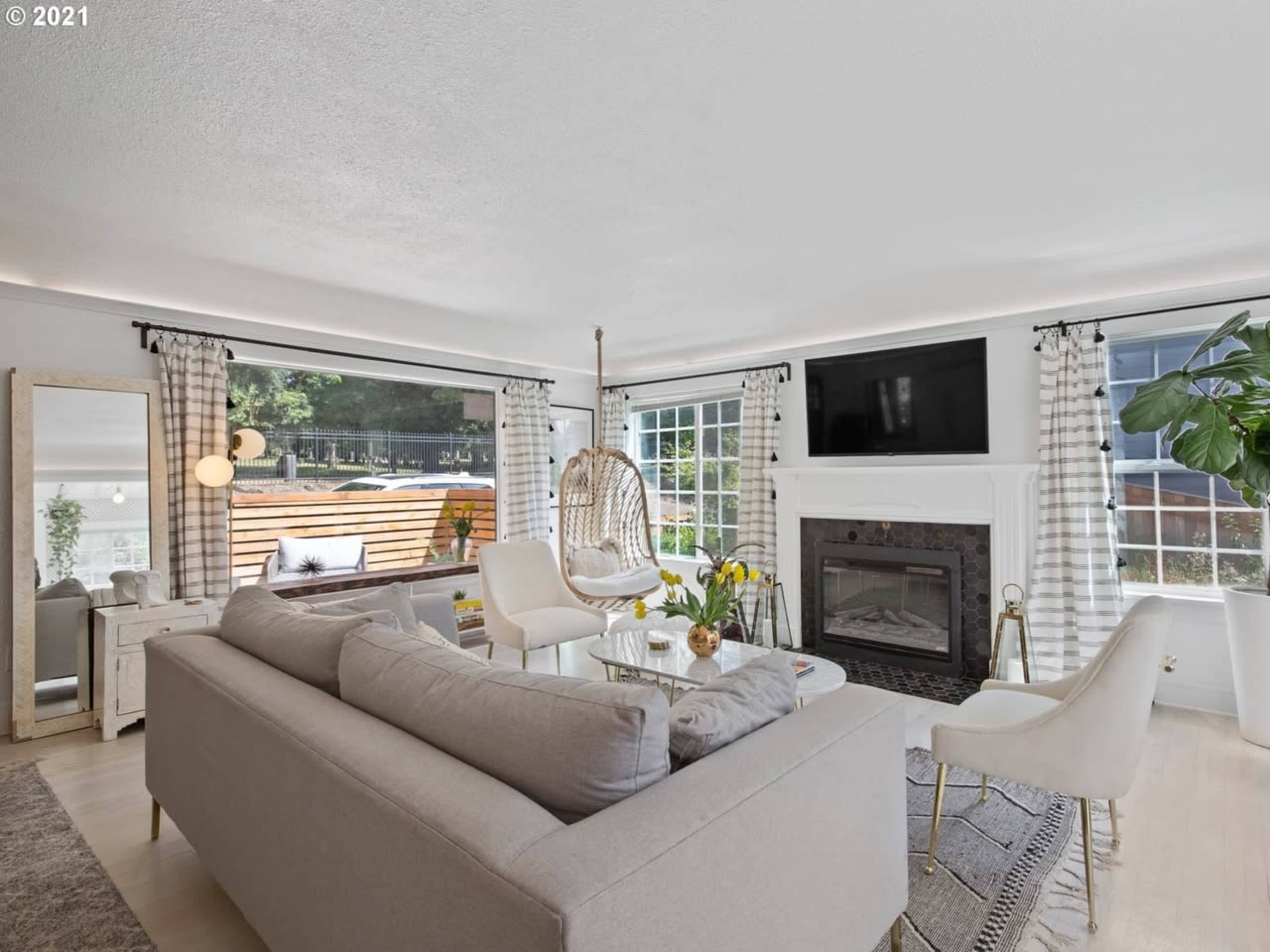 A well-lit living room featuring a light gray sofa, a hanging chair, a fireplace, and large windows with sheer curtains.