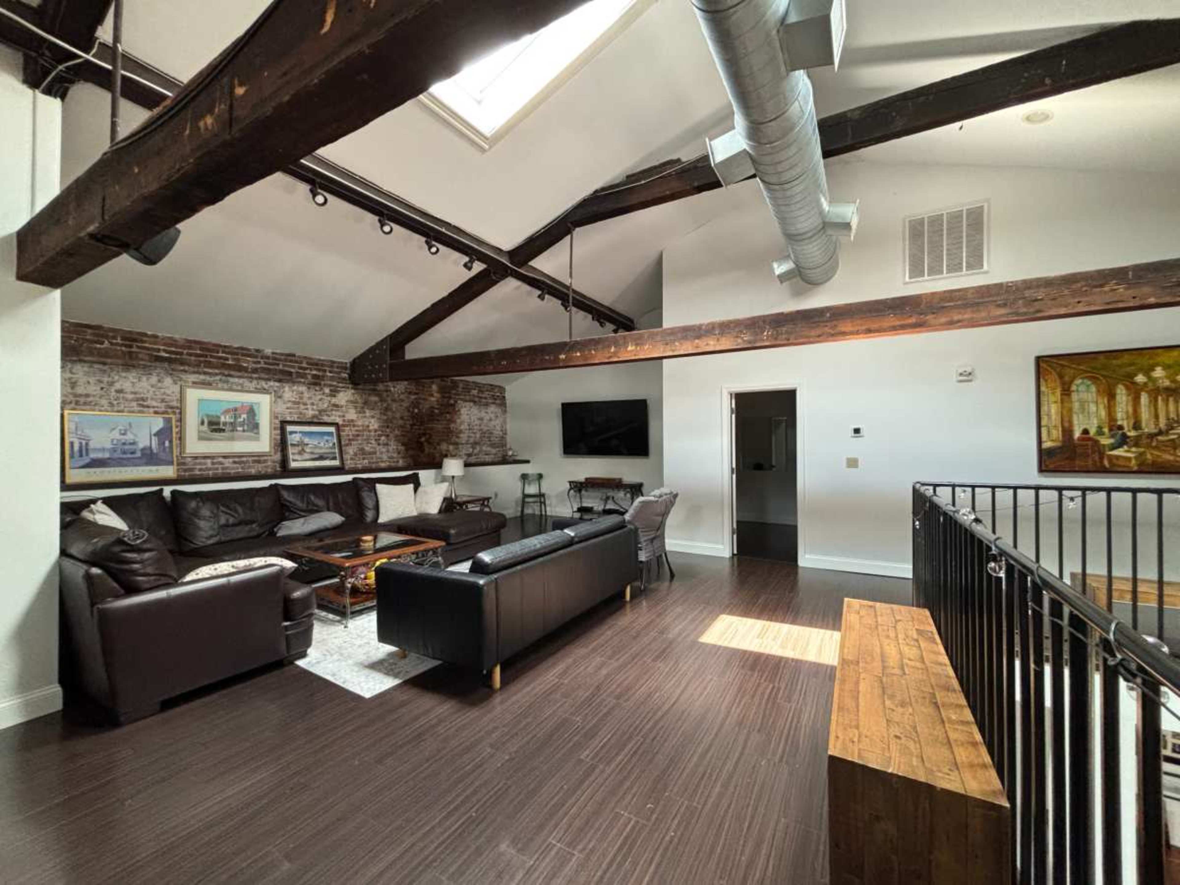The image shows a spacious living room with exposed brick walls, hardwood floors, and a high ceiling featuring wooden beams.