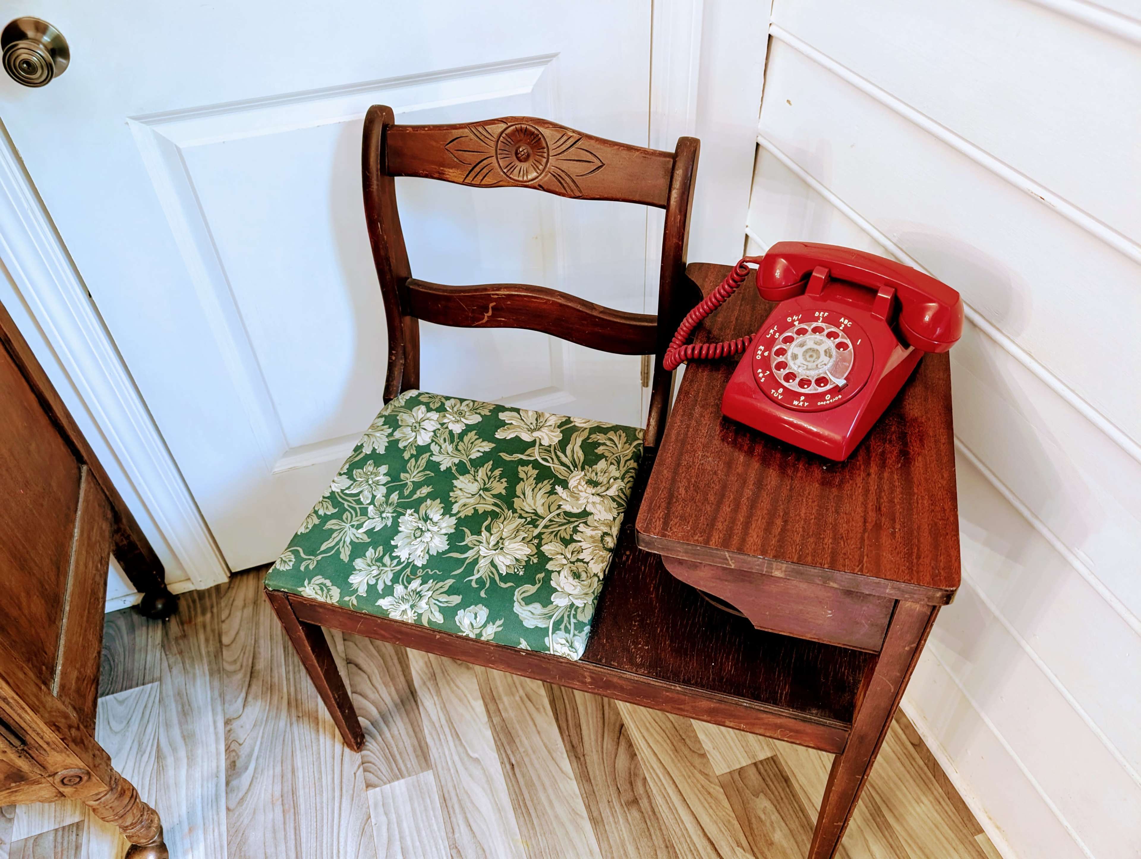 A vintage wooden chair with a floral cushion sits next to a small wooden table topped with a red rotary phone.