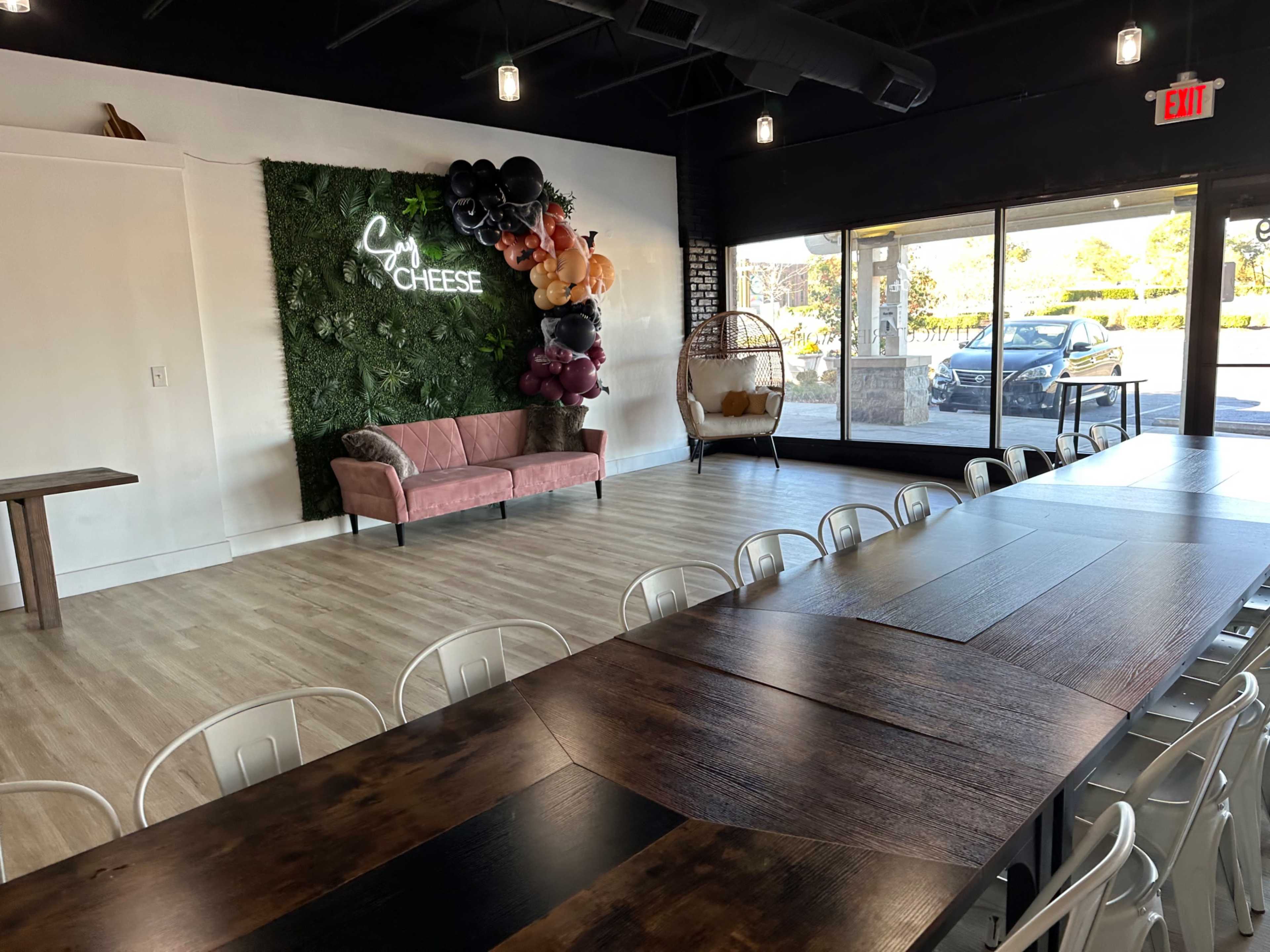 The interior of a café features a long wooden table, white chairs, a pink couch, and a decorative green wall with a sign that reads "Say Cheese."