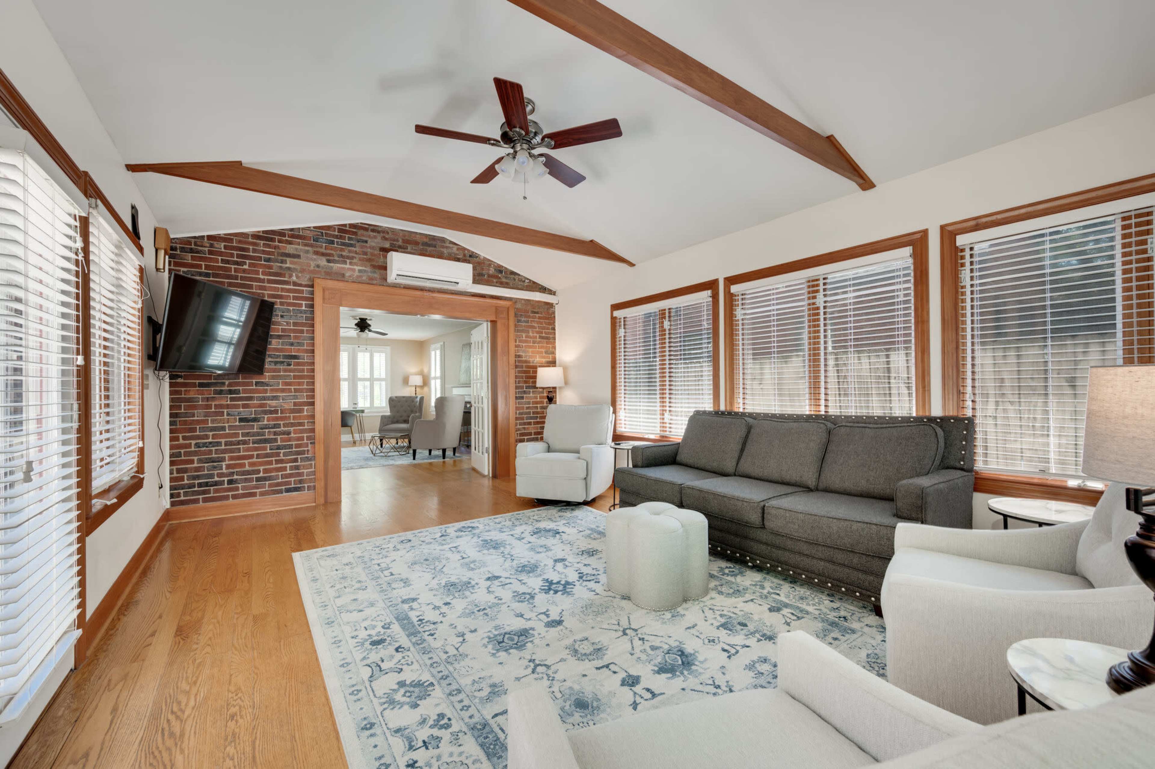 The living room features a gray sofa, two armchairs, a coffee table, a television mounted on the wall, and large windows with blinds, creating a bright and open atmosphere.