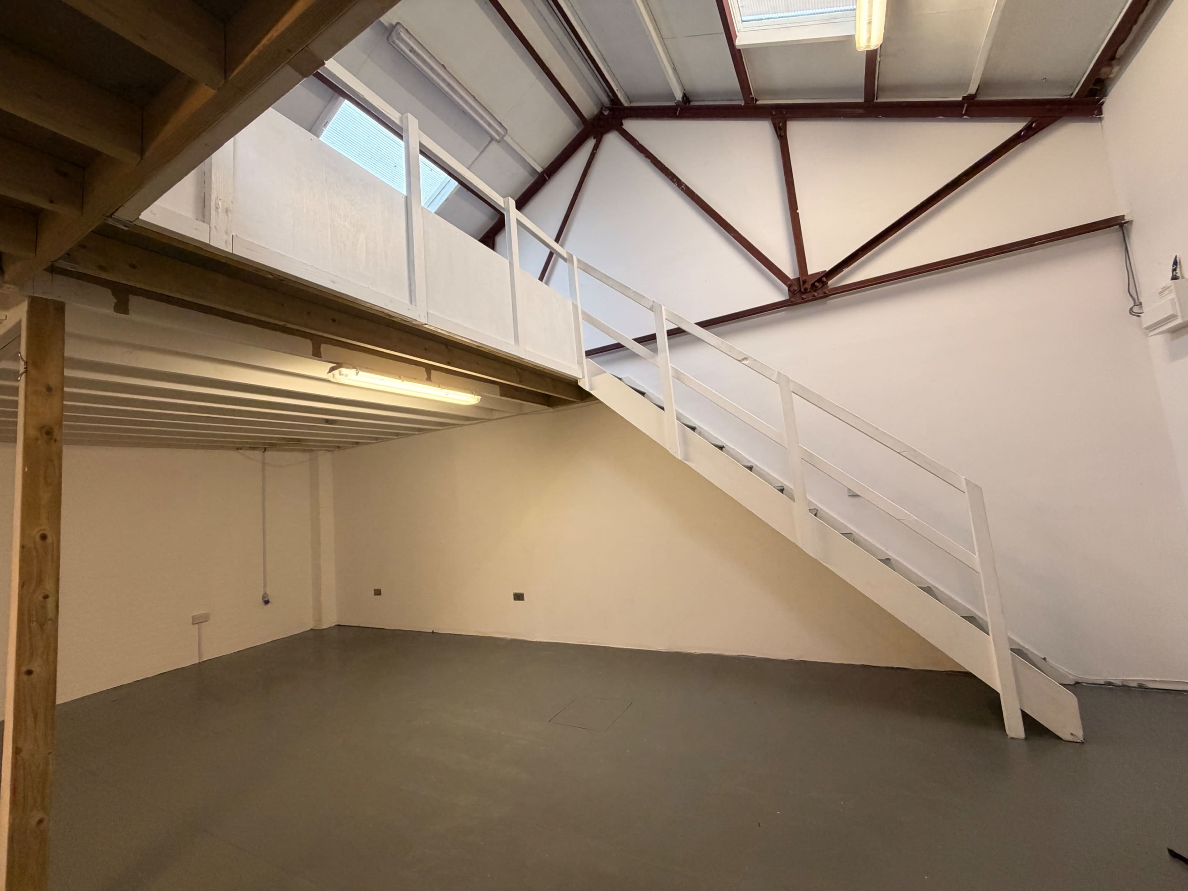 The image shows an empty room with a white staircase leading to a loft area and exposed metal beams on the ceiling.