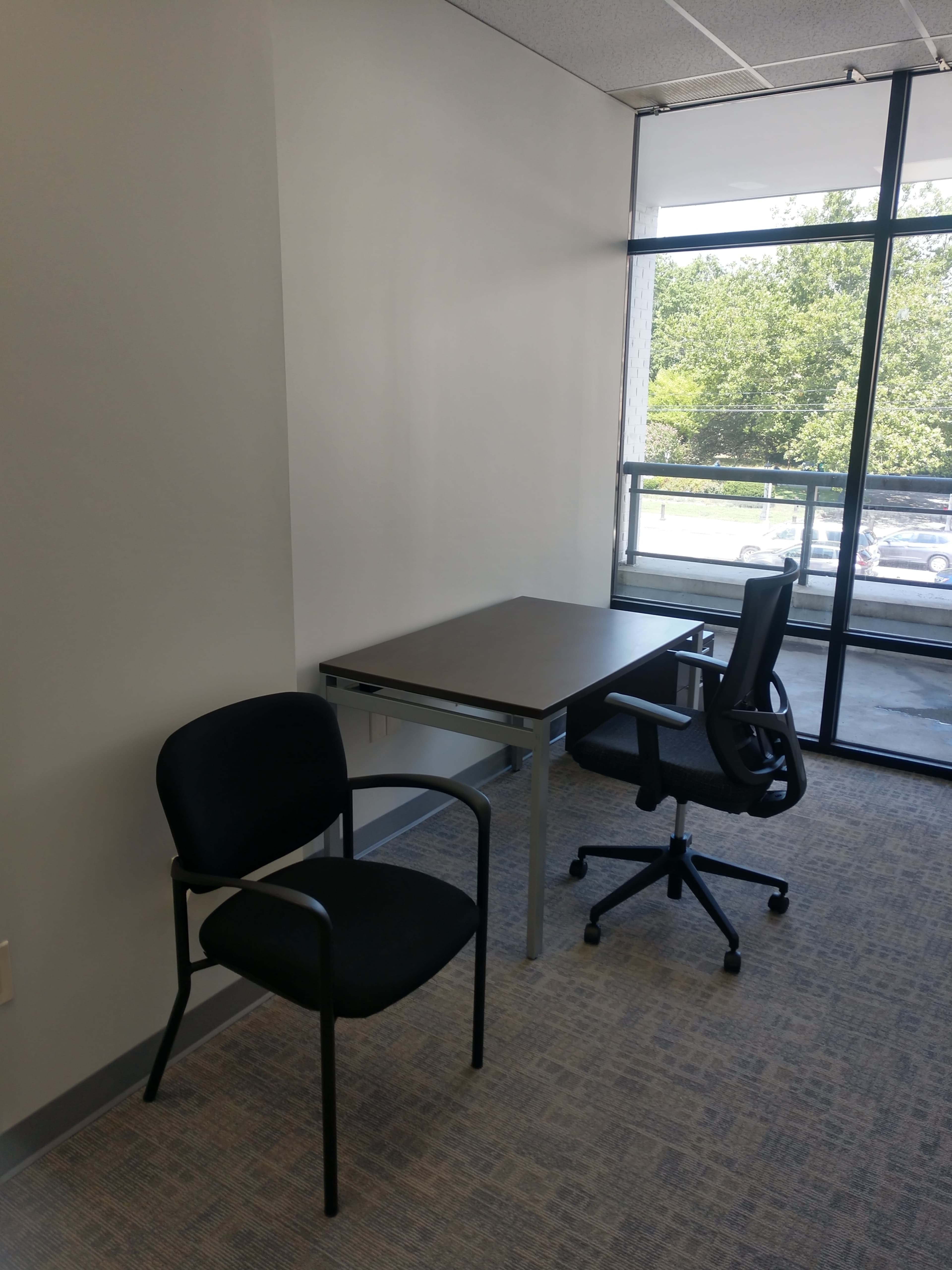 The image shows a small office space with a table and two chairs, situated near a large window overlooking a green landscape.