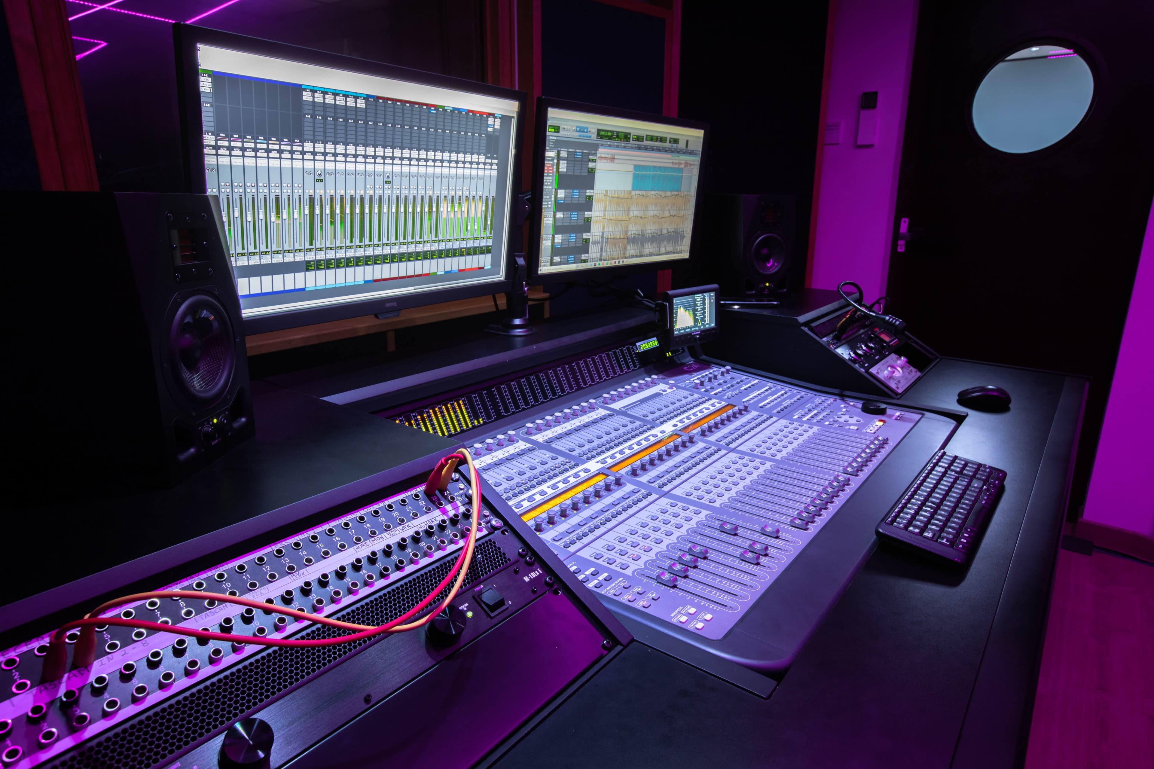 A recording studio with a large mixing console, computer monitors displaying digital audio software, and various audio equipment is illuminated by purple lighting.