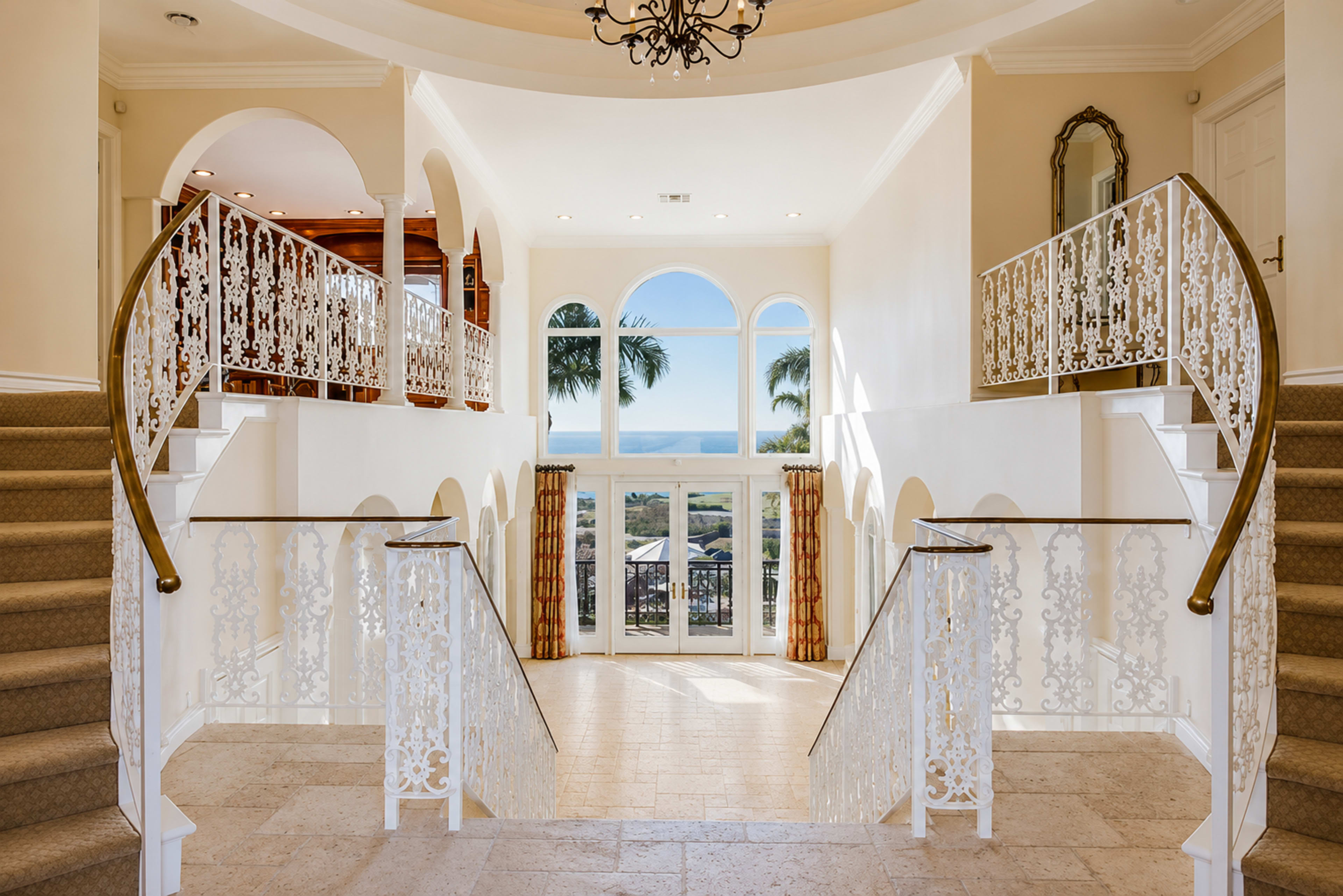 The image shows a grand entrance hall with a double staircase, adorned with wrought iron railings, leading to a set of double doors that open to a view of the outdoors.