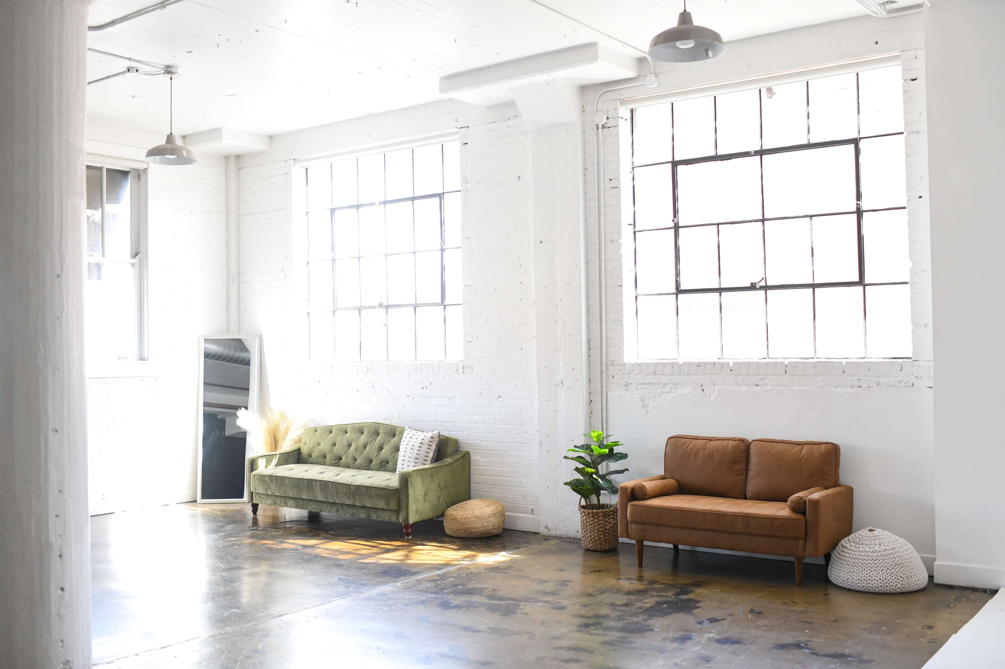 A bright, modern room with large windows, featuring a green velvet sofa, a brown couch, and a potted plant.