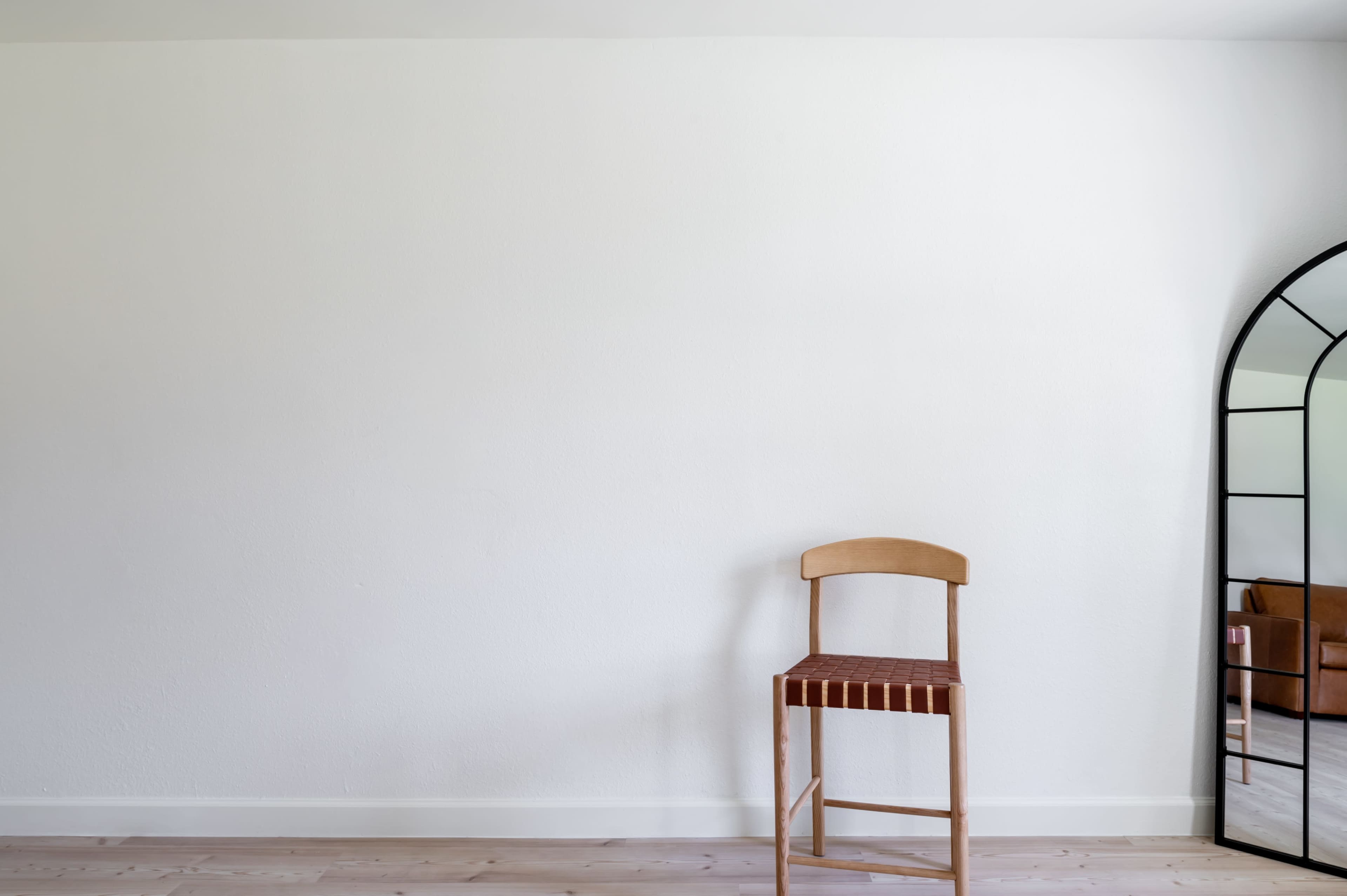 A wooden stool stands beside a tall, arched mirror against a white wall in a simple interior space.