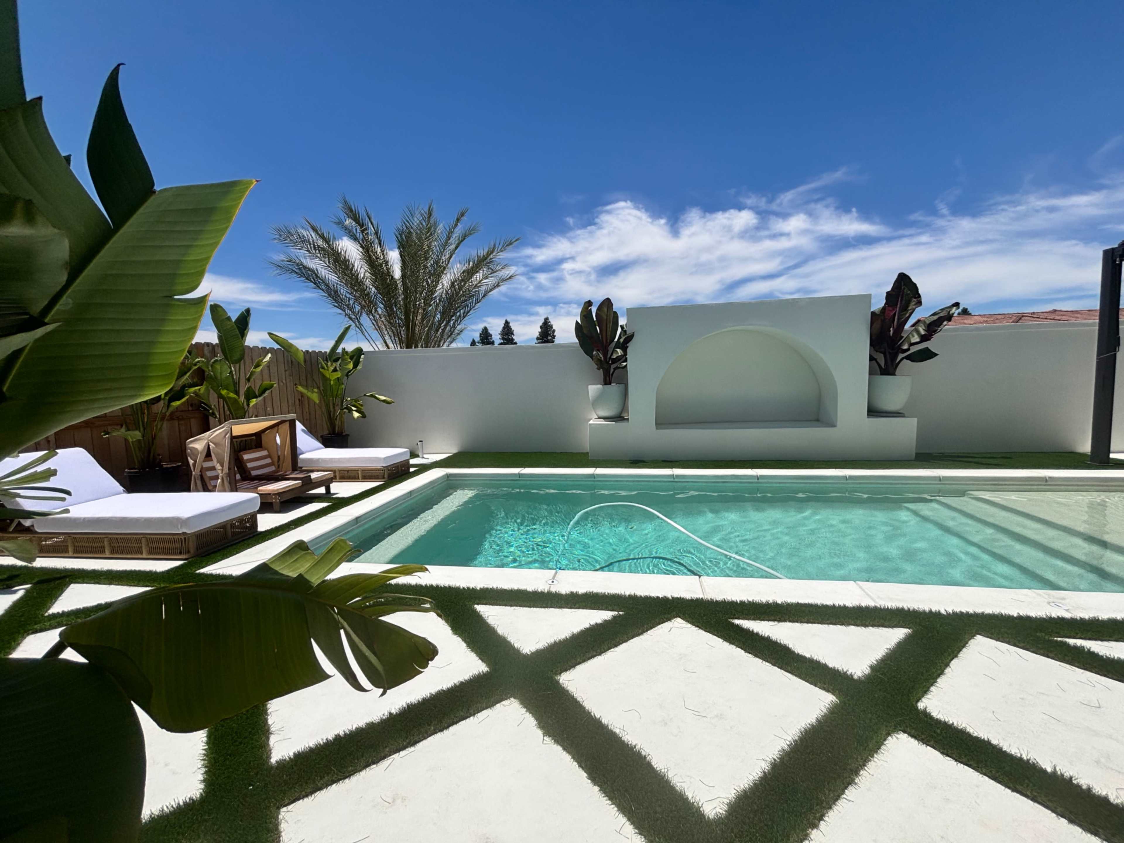 The image shows a pool area surrounded by white walls, featuring lounge chairs, a decorative wall, and tropical plants under a blue sky.