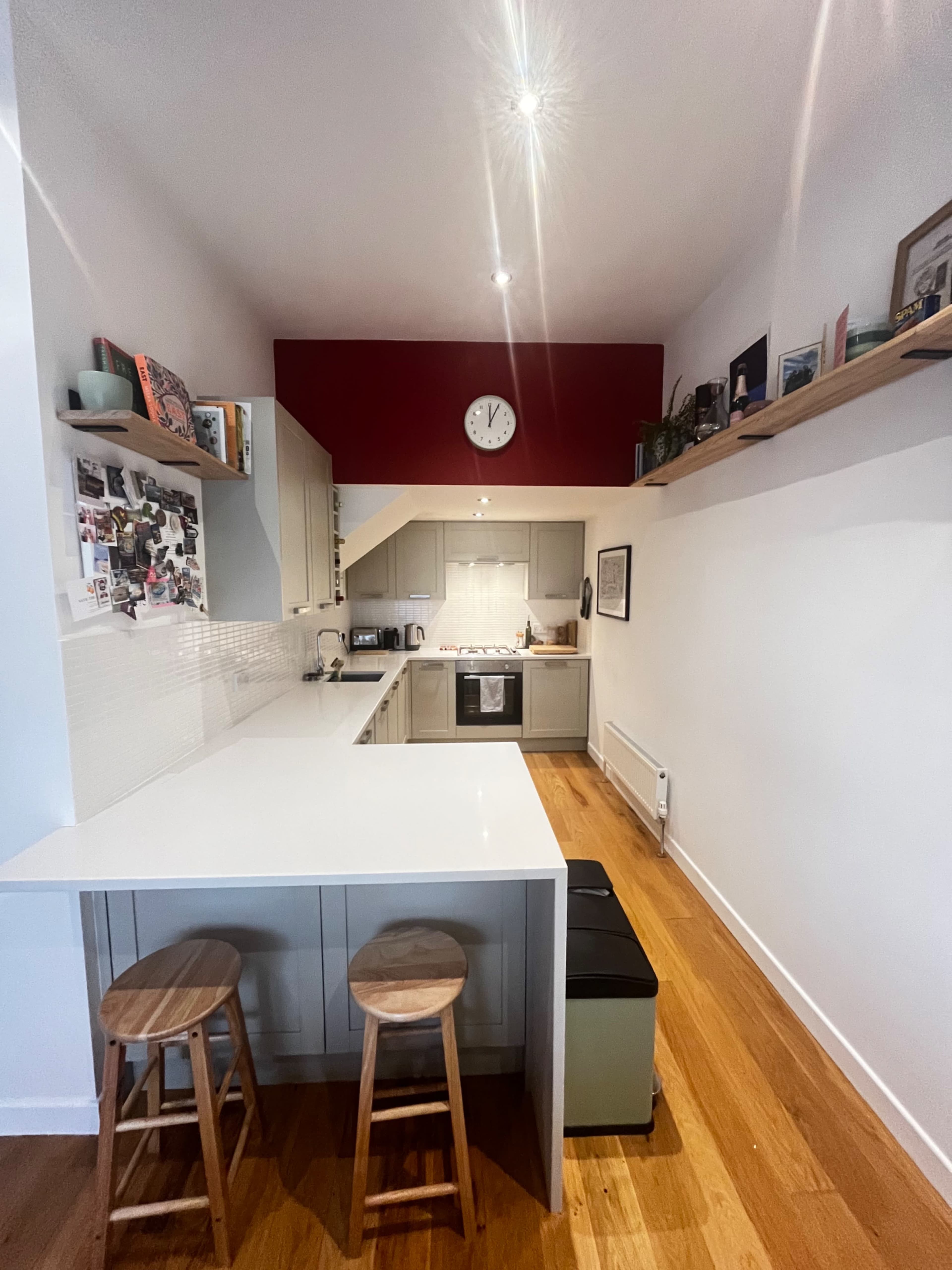 A modern kitchen features a white countertop, wooden stools, and a red accent wall with a clock.