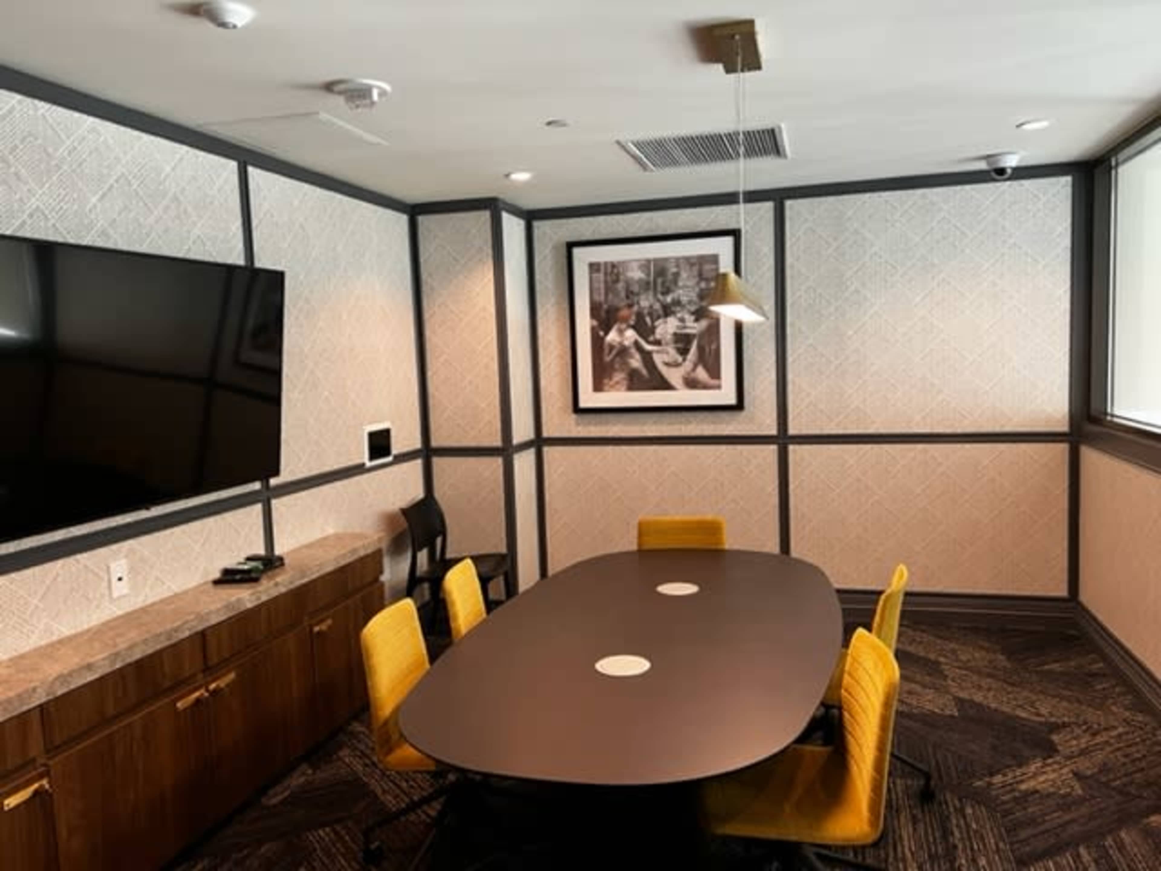 A conference room with an oval table, four yellow chairs, a large television on the wall, and a framed black-and-white photograph.