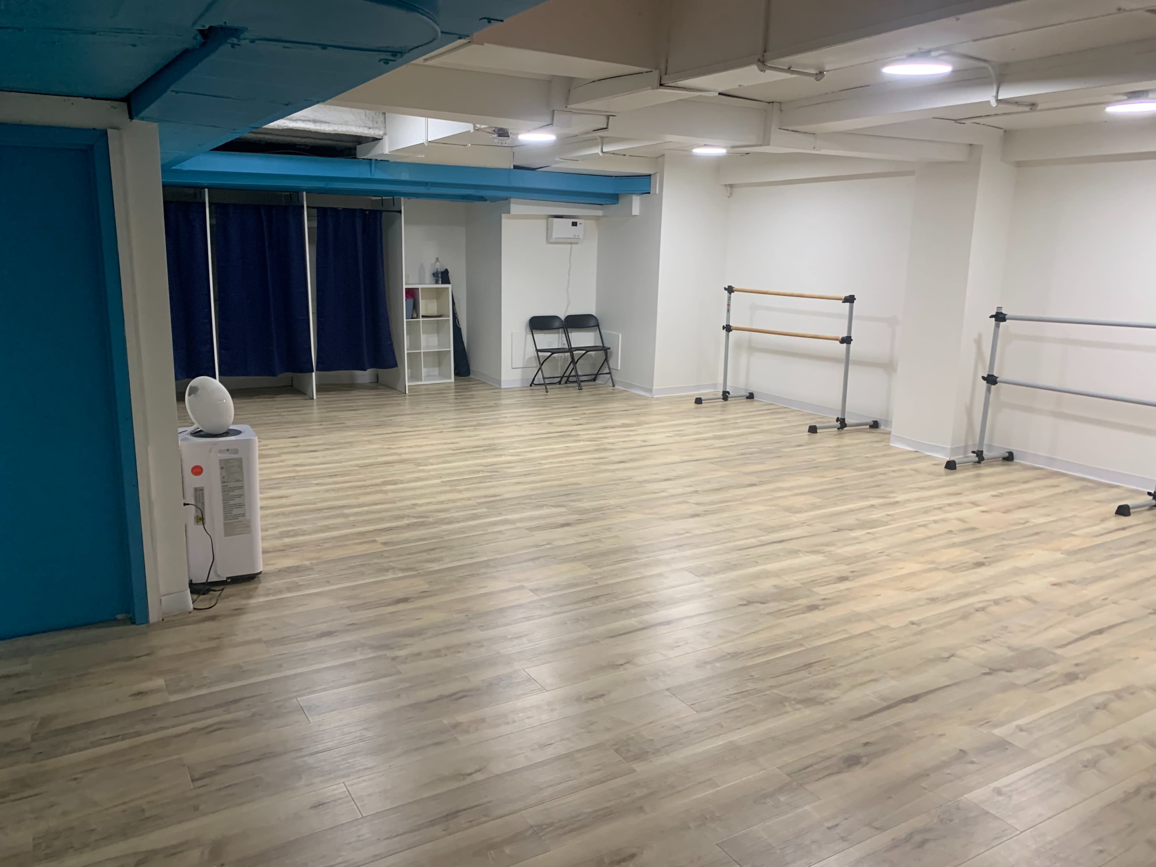 The image shows a spacious, empty practice studio with light wooden floors, blue and white walls, and mirrored ballet bars along one side.