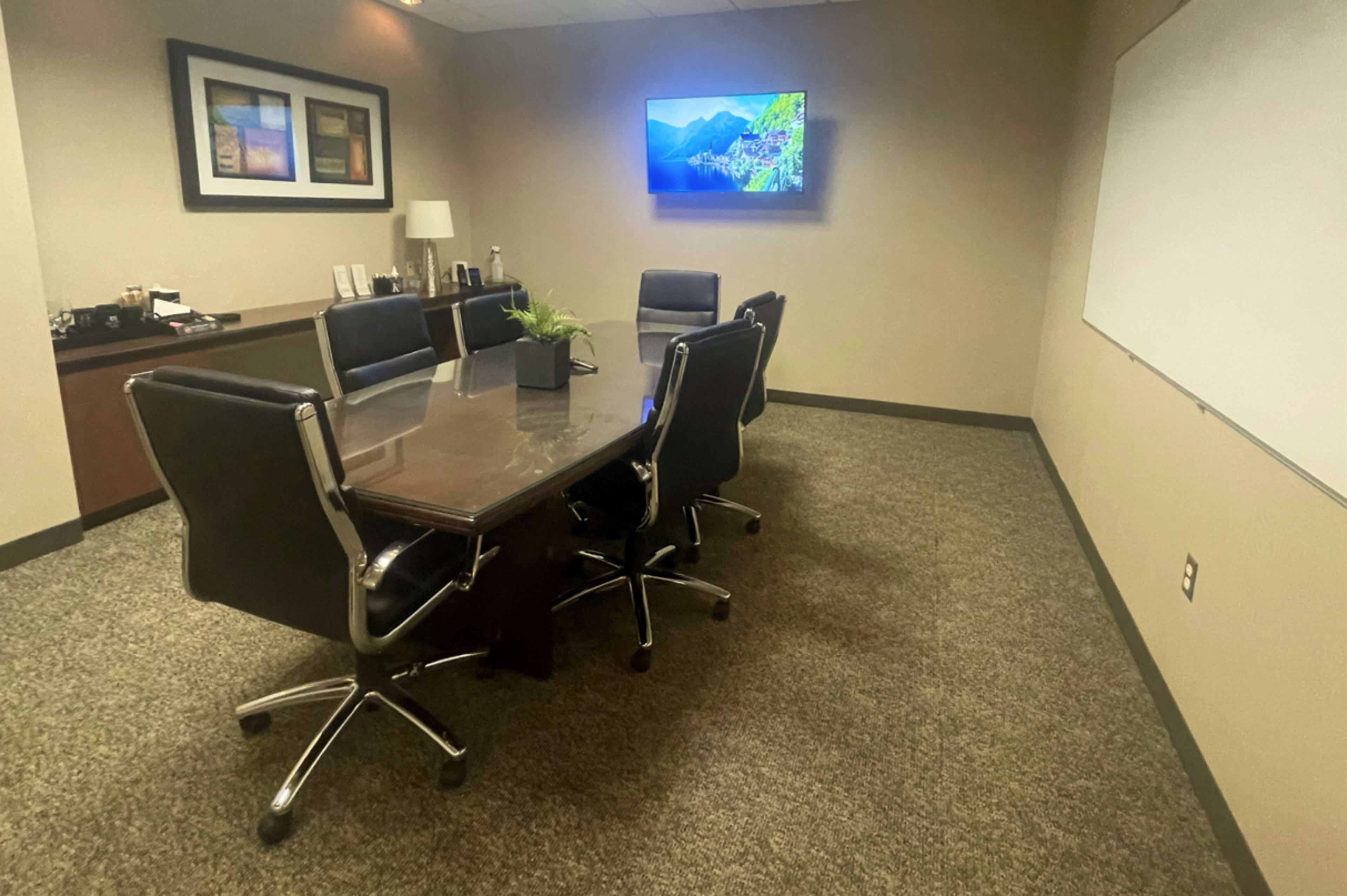 A small conference room features a glass table surrounded by four black chairs, a television on the wall, and a whiteboard in the corner.