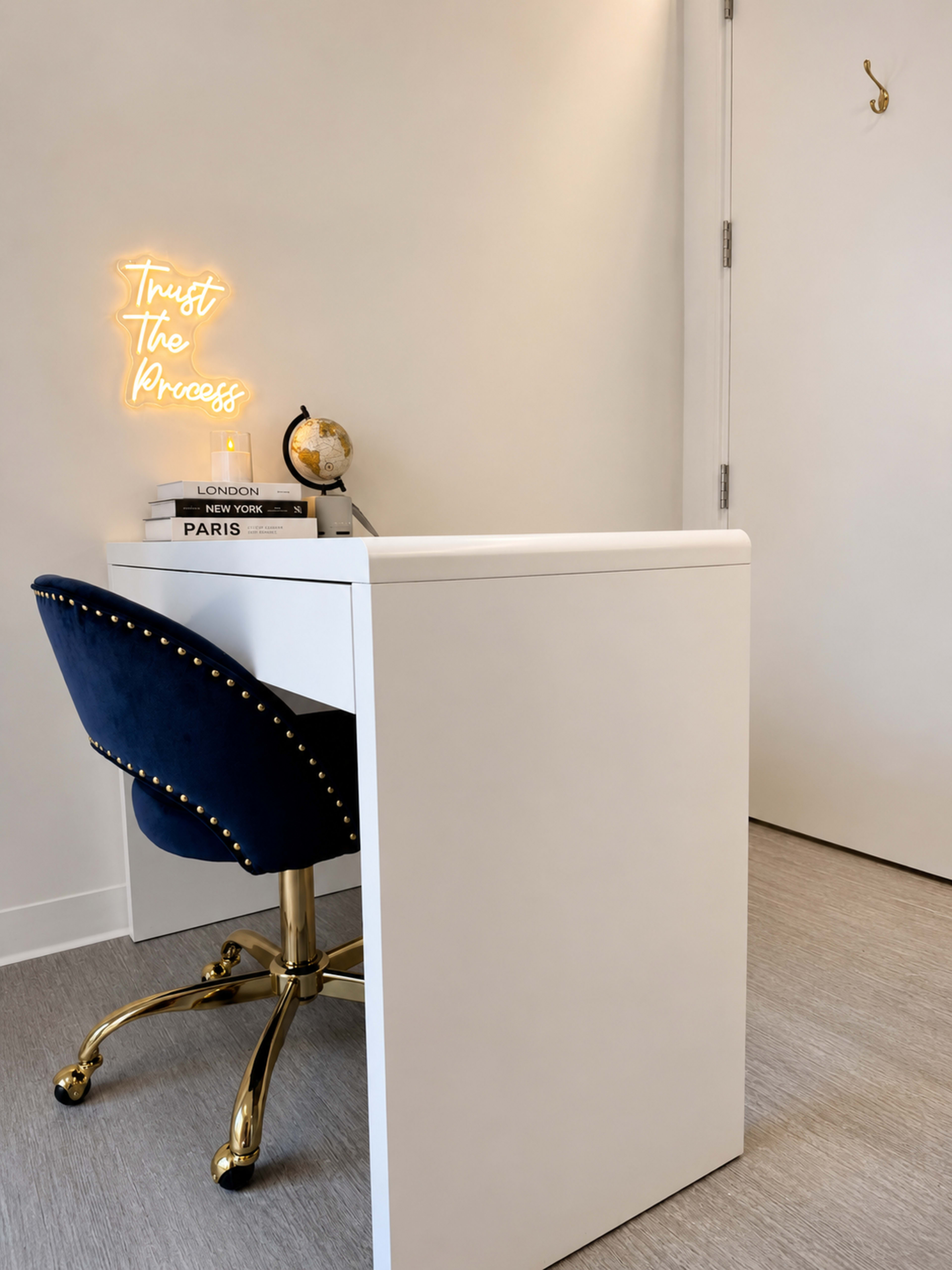 A modern desk with a blue upholstered chair features a neon sign that reads "Trust The Process" and a decorative globe, set against a simple white wall.