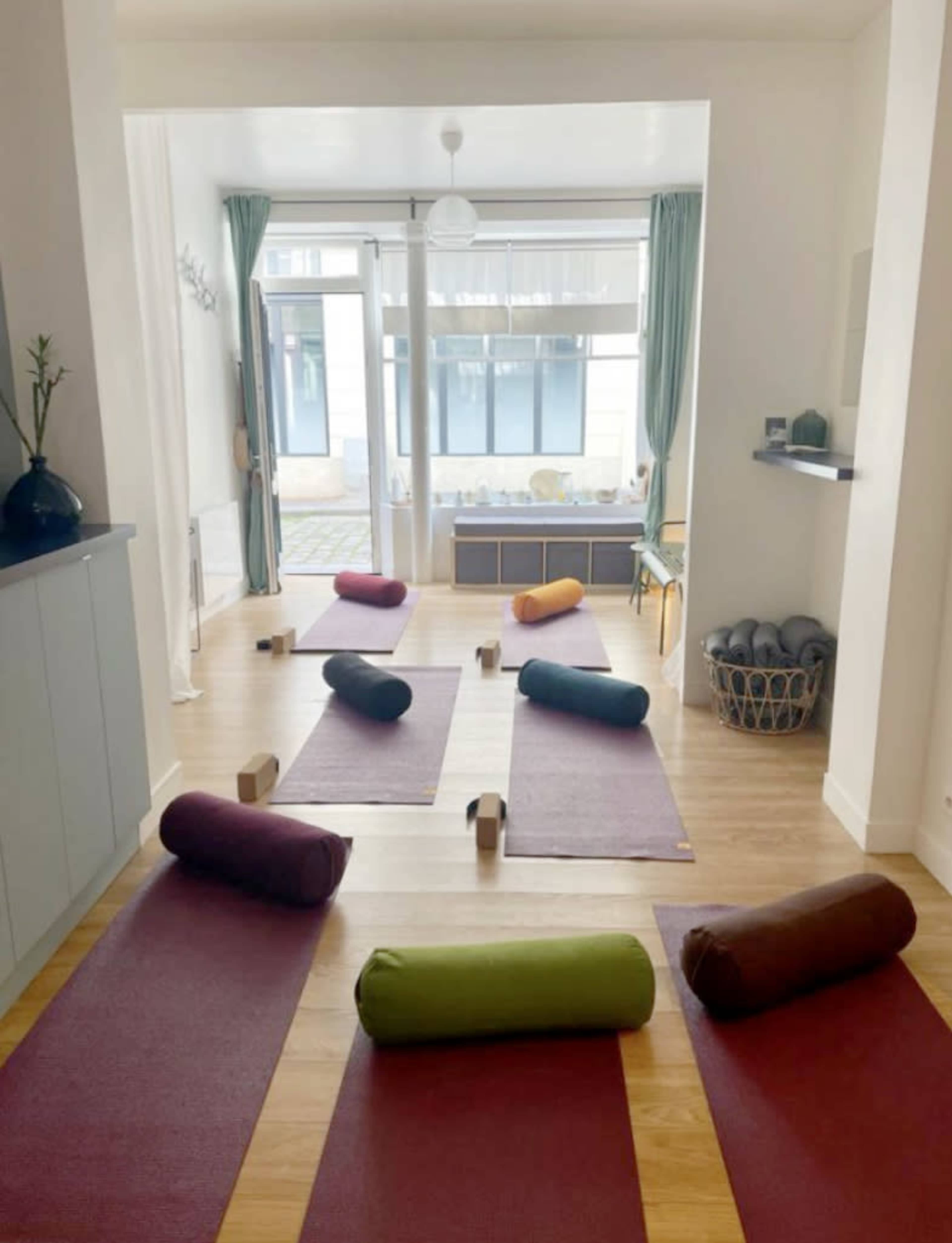 A spacious yoga studio features several exercise mats and bolster cushions arranged on a wooden floor, with large windows allowing natural light to fill the room.