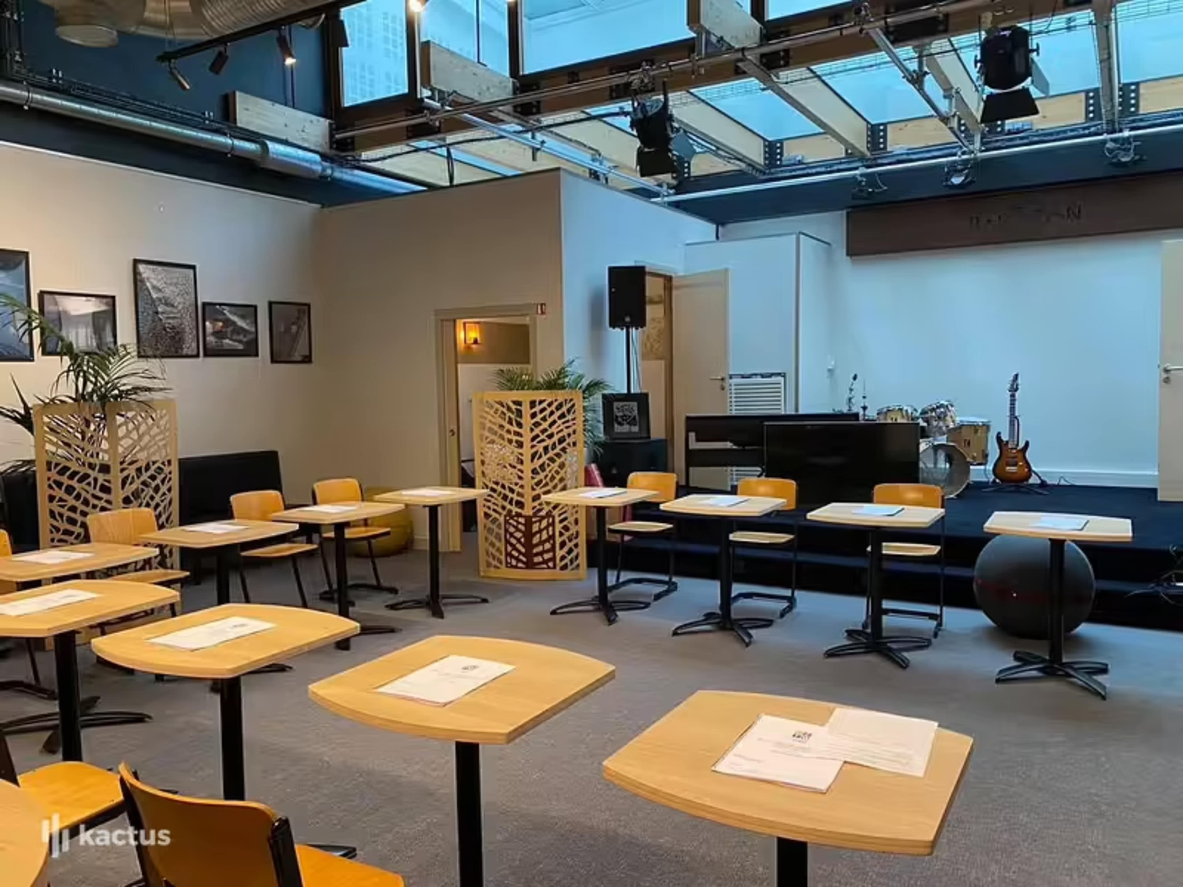 A modern seminar room with rows of wooden tables and chairs facing a small stage with musical instruments in the background.