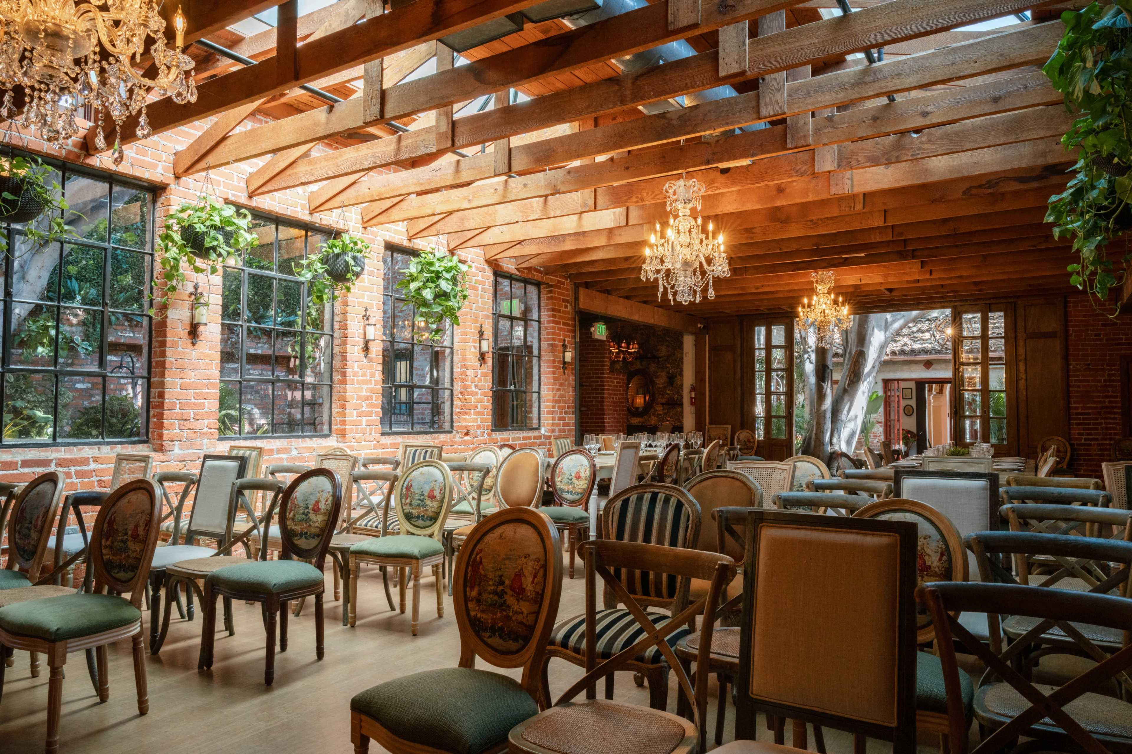 The image shows a spacious indoor venue with wooden beams, brick walls, and elegantly arranged chairs adorned with decorative upholstery.