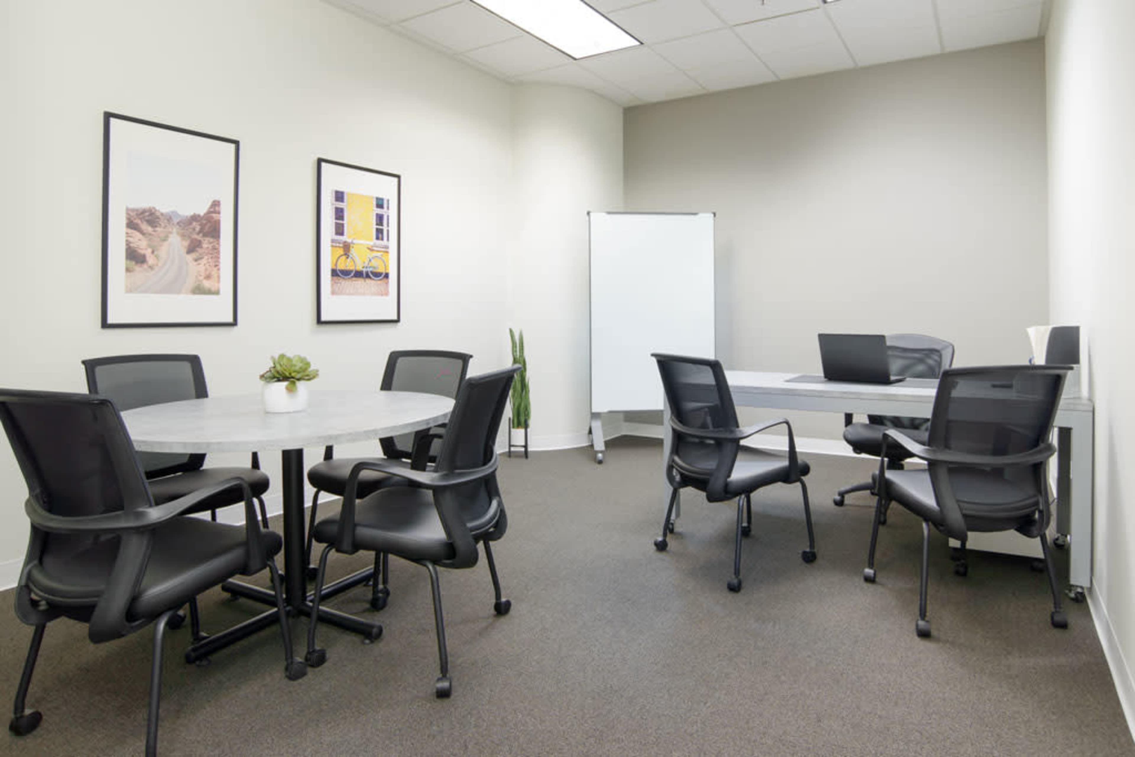 A minimalistic meeting room with a round table, six black chairs, a whiteboard, and a laptop on a rectangular table.