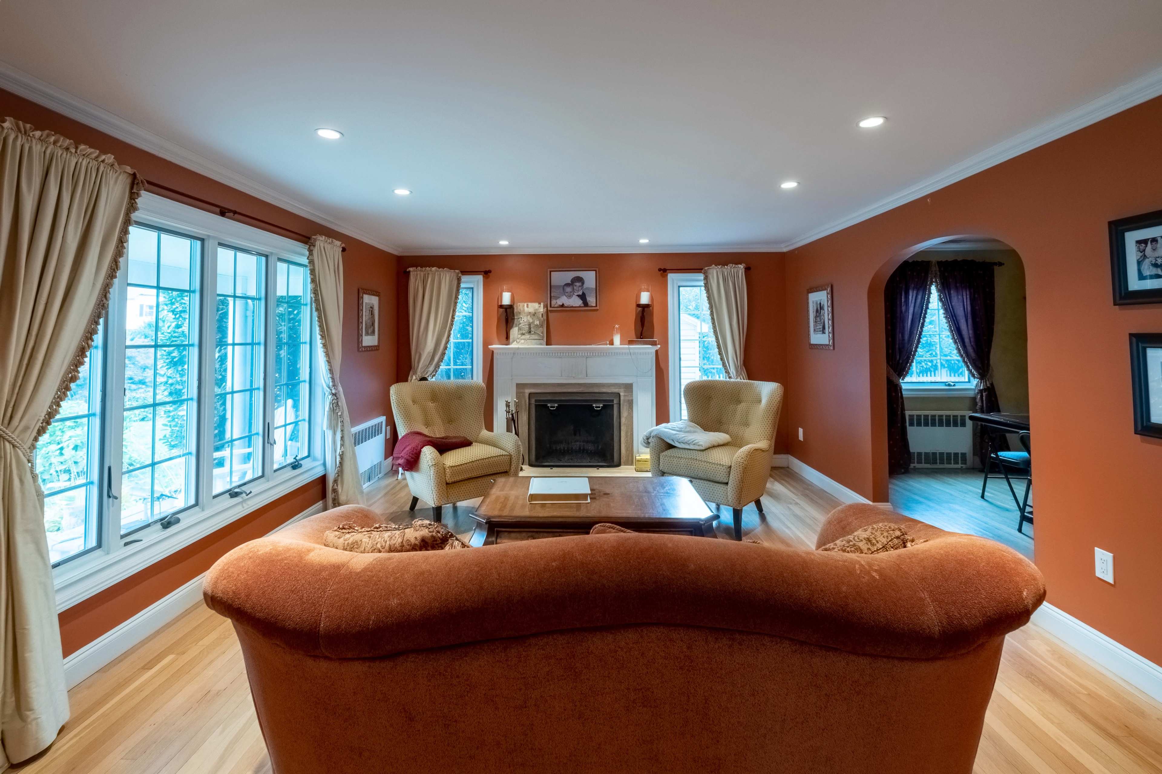 A cozy living room features an orange wall, a fireplace, two armchairs, and large windows allowing natural light.