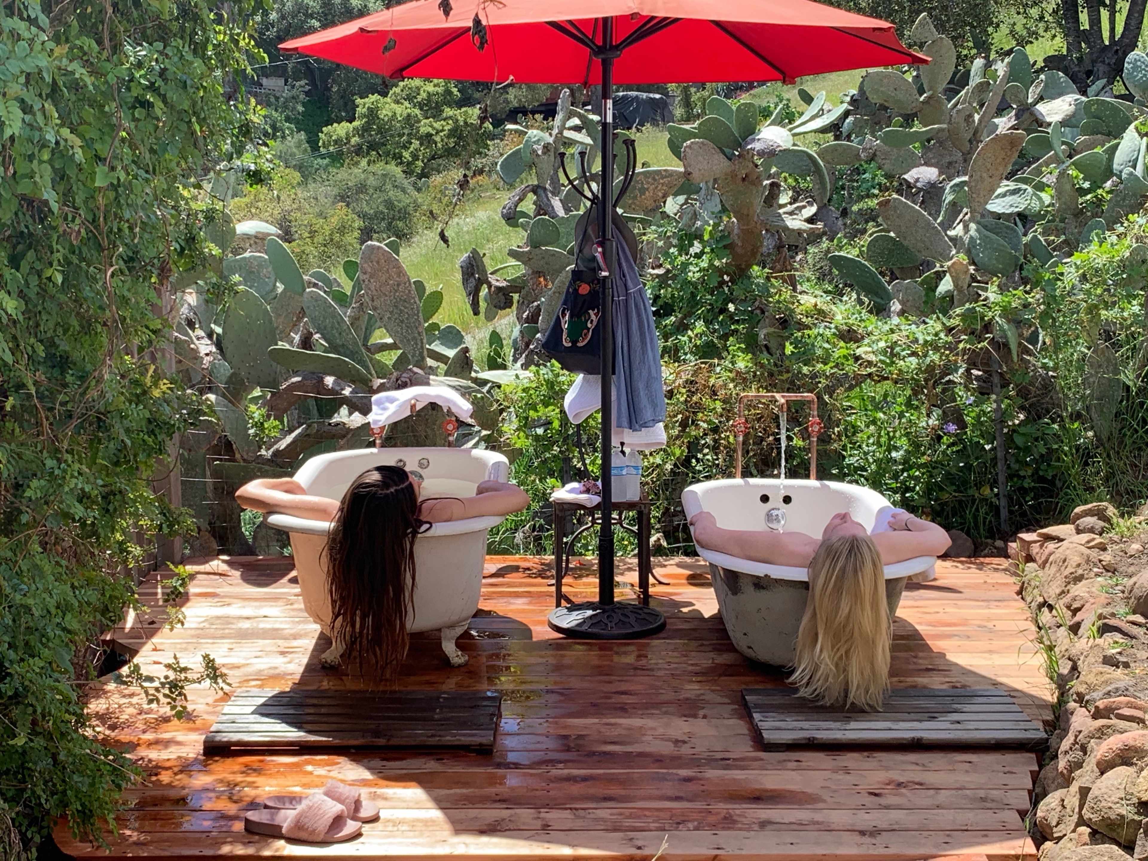 Two bathtubs are positioned on a wooden platform under a red umbrella, surrounded by lush greenery and cactus plants.