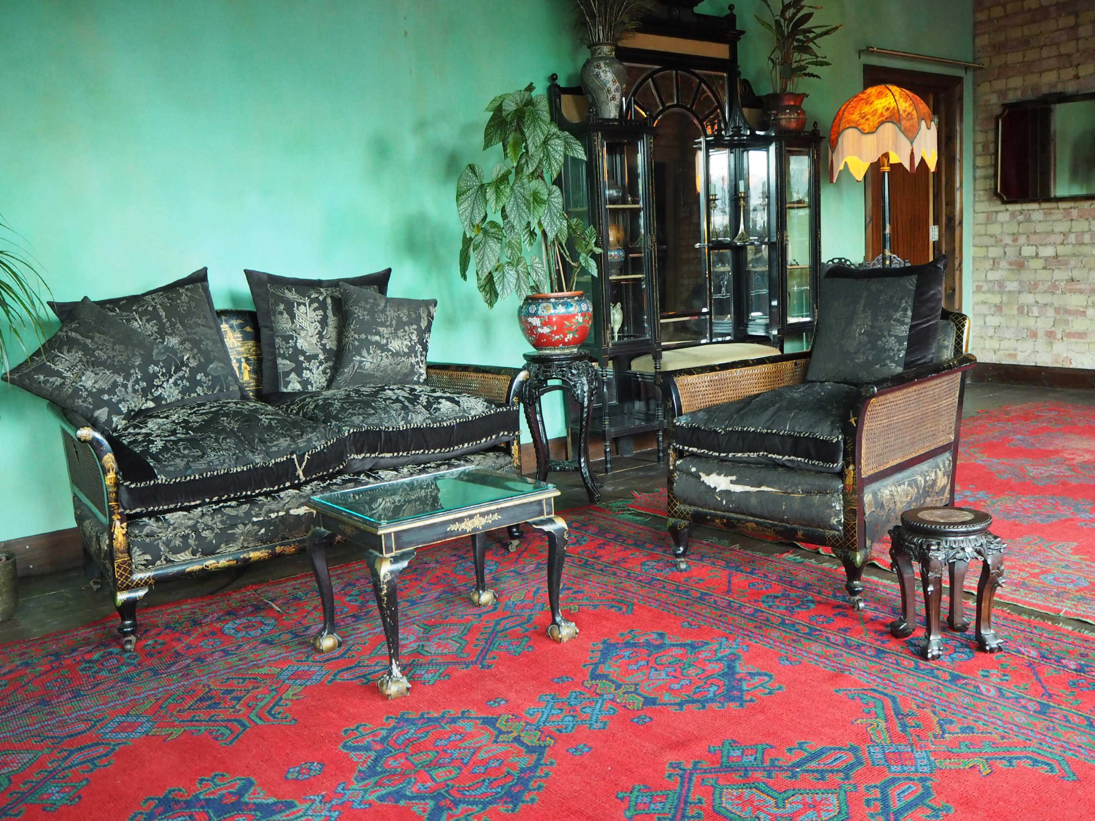 A vintage living room with a dark upholstered sofa and armchair, a glass coffee table, and a patterned red rug, under soft lighting from a lamp.