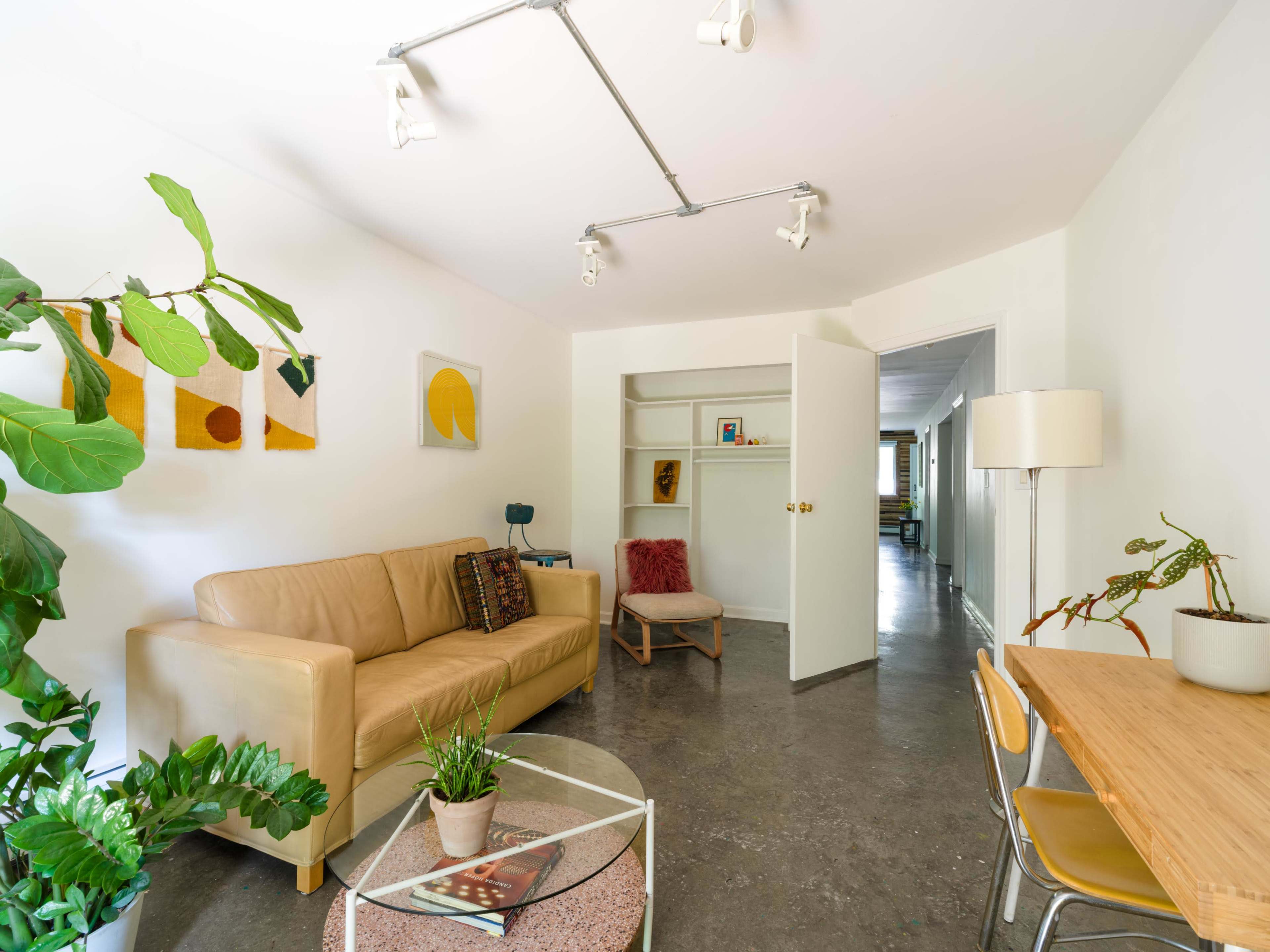 The image shows a bright, modern living room with a beige couch, a glass coffee table, a small dining table, and minimalist decor.