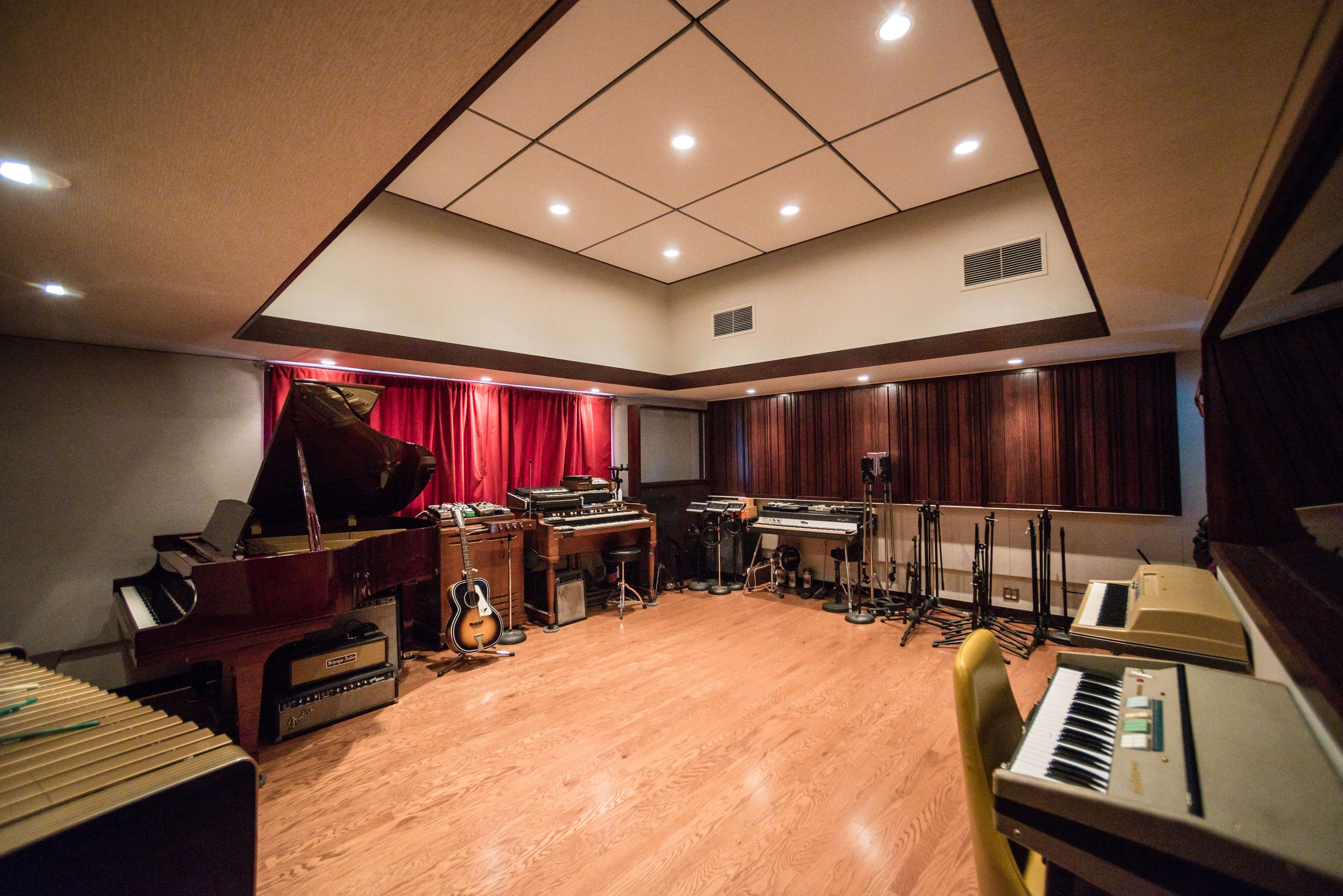 The image shows a music studio with various instruments, including a grand piano, guitars, and synthesizers, arranged within a room featuring wooden walls and red drapes.