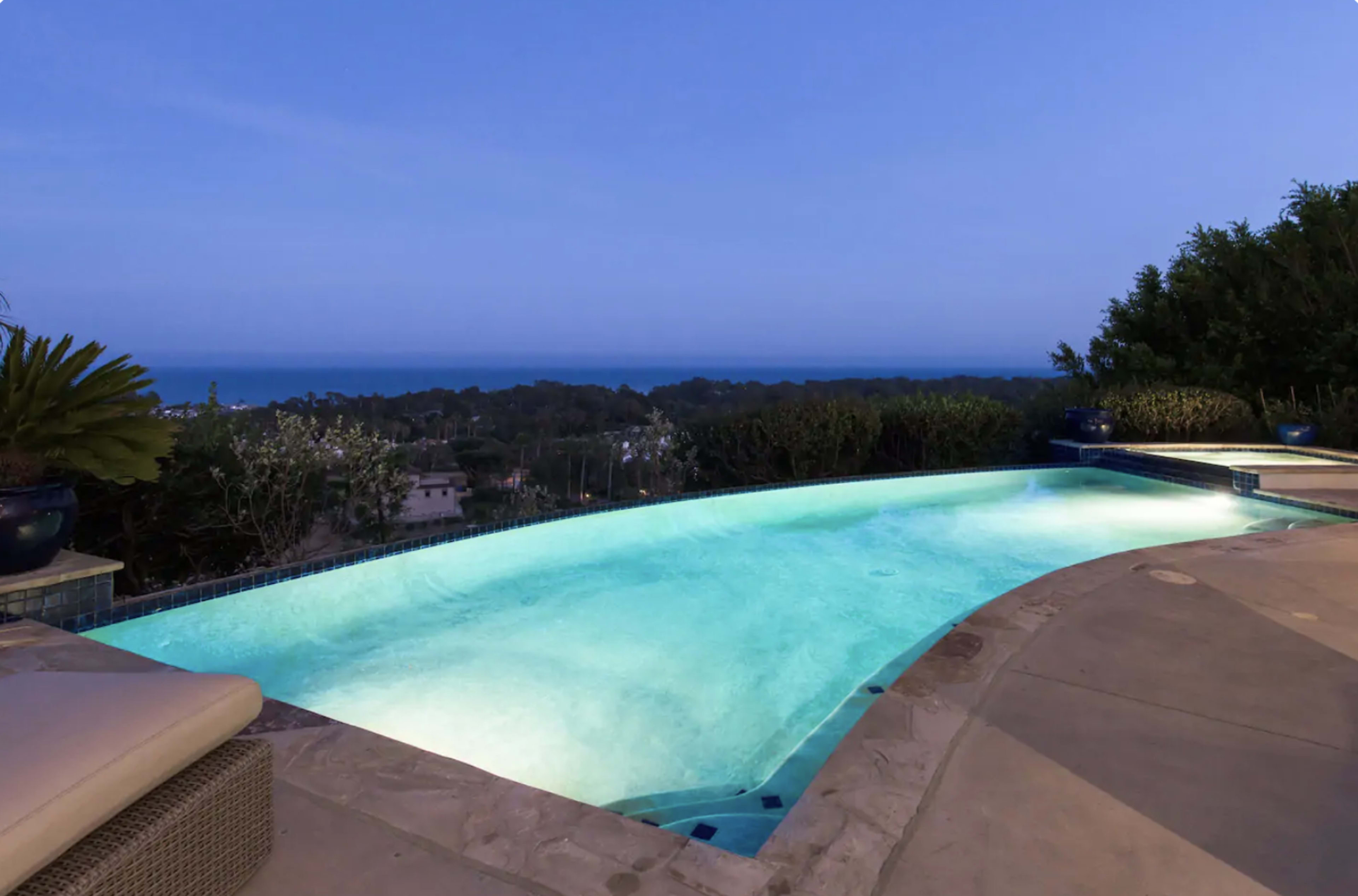A glowing swimming pool overlooks a coastal landscape at dusk.