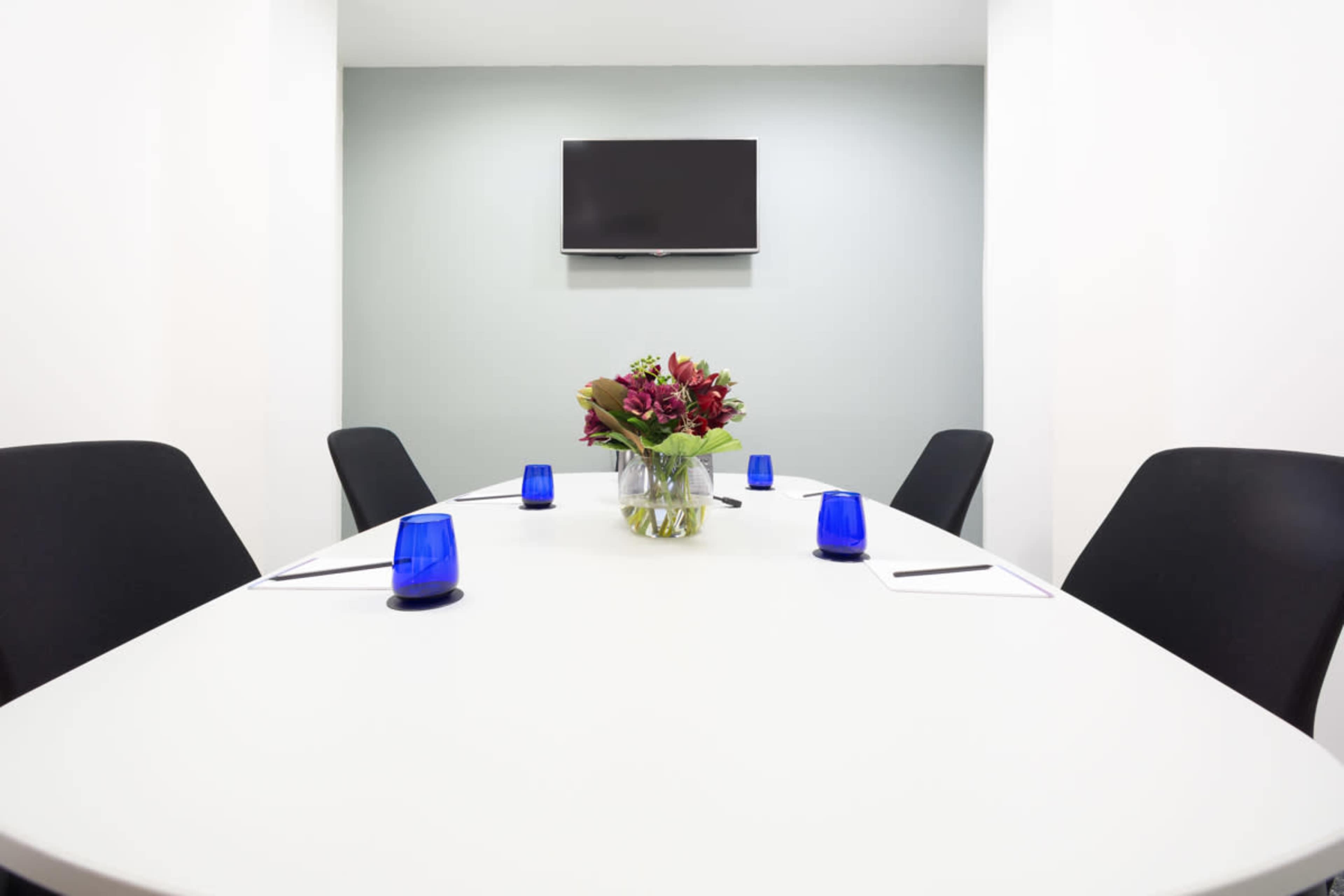 A conference room features a white oval table with a floral centerpiece, surrounded by black chairs, and a wall-mounted television.