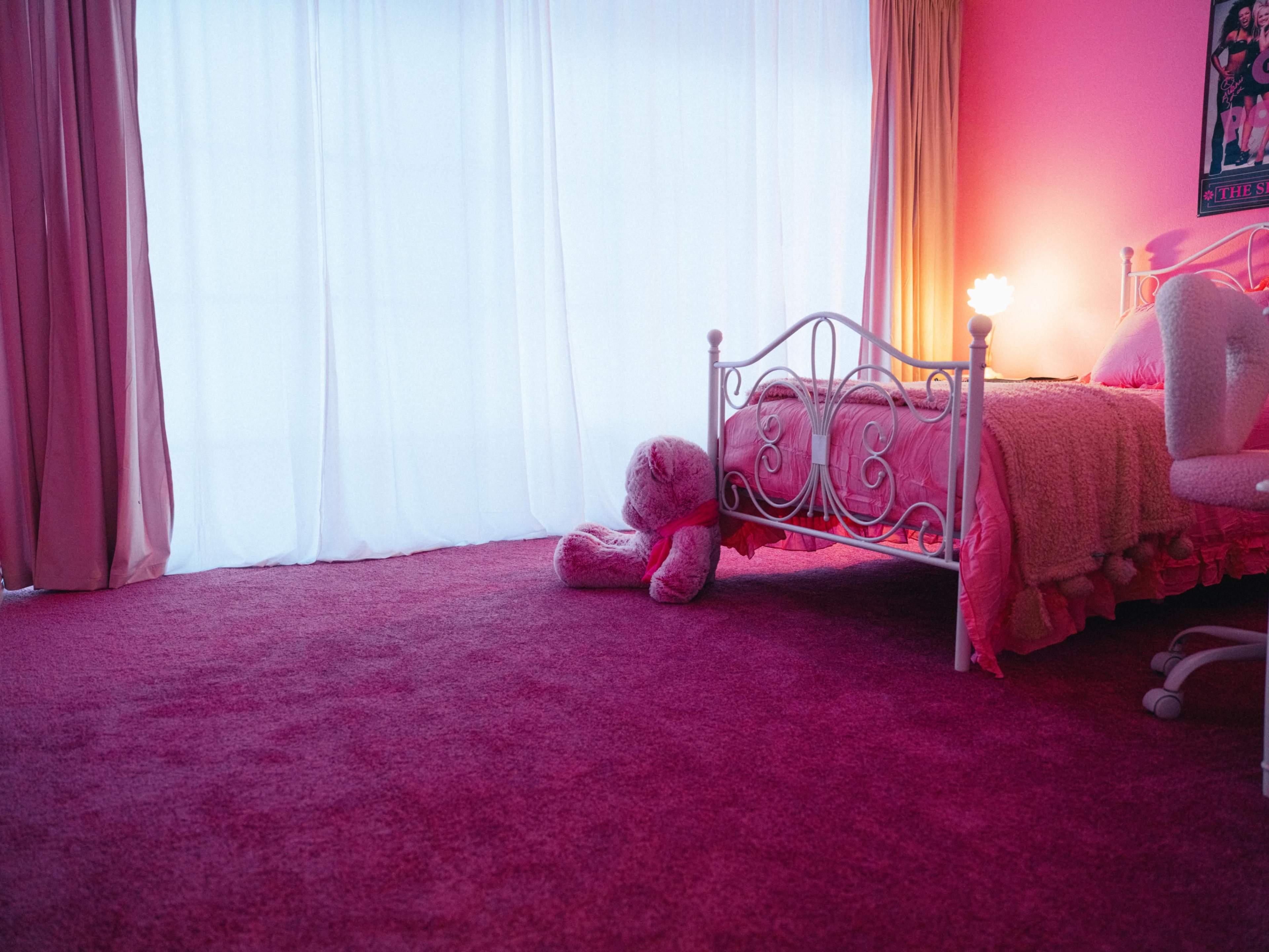 A pink-themed bedroom features a bed with a plush blanket, a large stuffed teddy bear on the carpet, and a soft glow from a lamp near the window.
