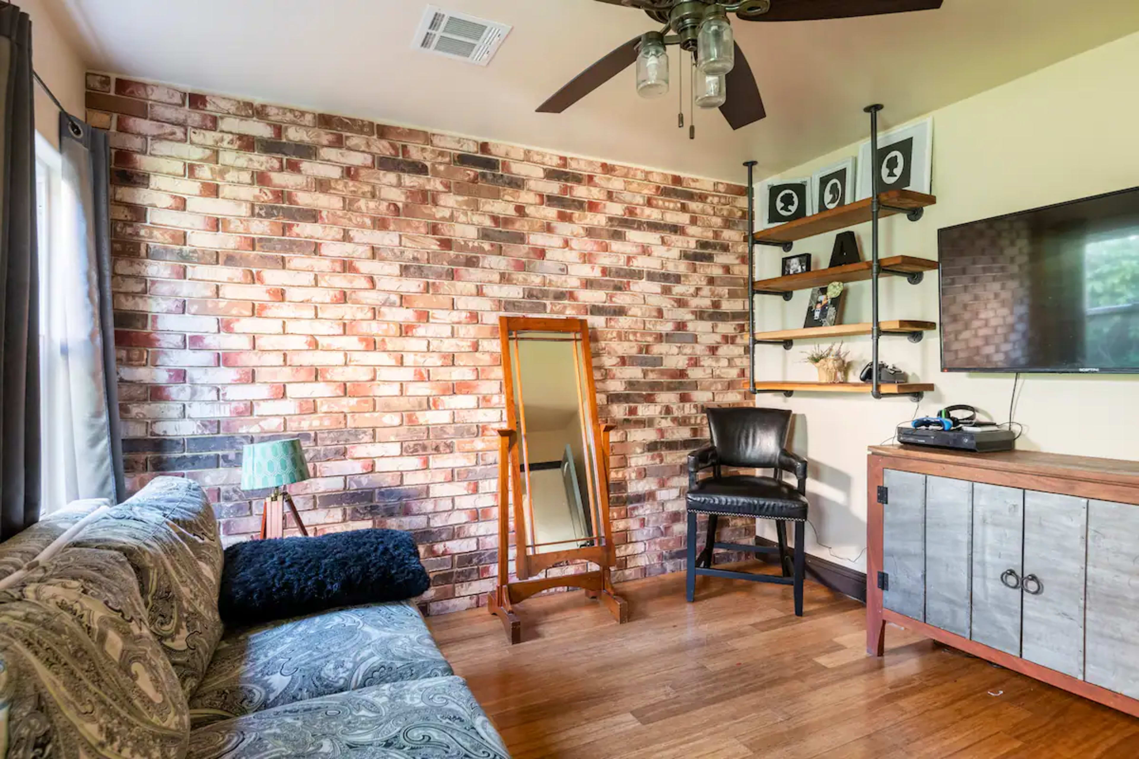 The room features a brick wall, a wooden chair, a full-length mirror, a television on a cabinet, and a patterned couch.