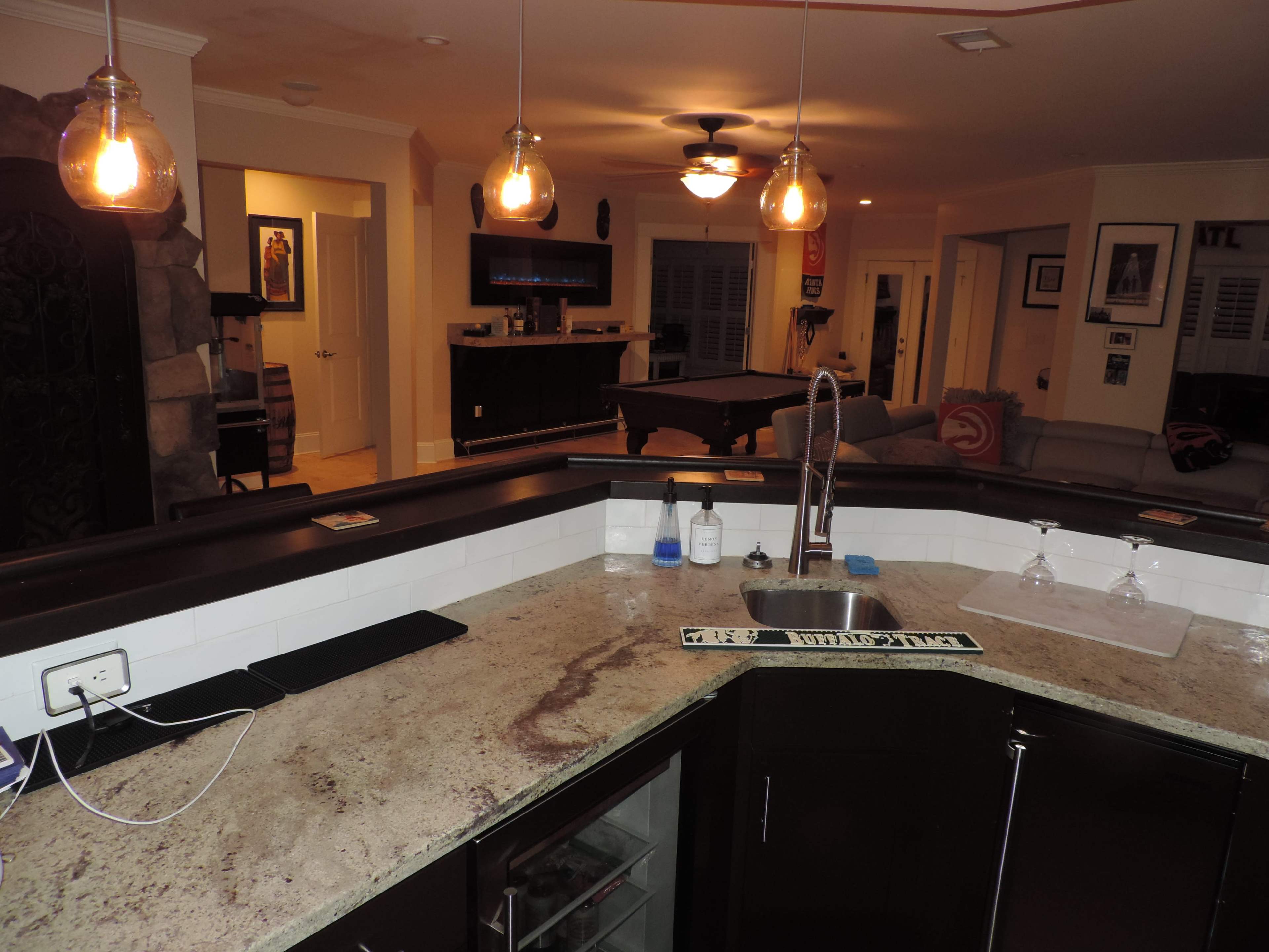 The image shows a home interior featuring a dark granite bar countertop with a sink, pendant lighting above, and an open living area with a pool table and seating in the background.